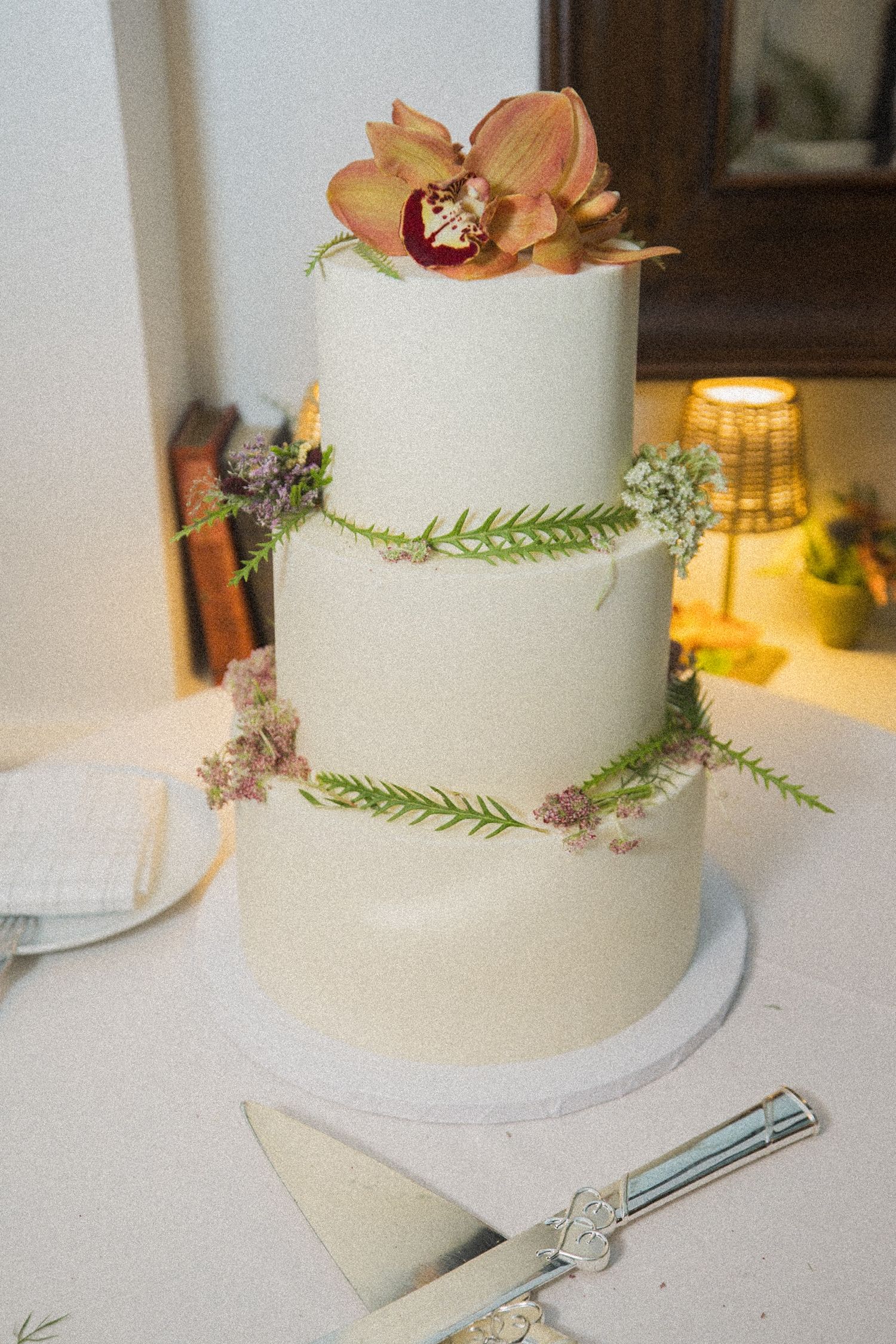 Three-tier white wedding cake decorated with greenery, small blossoms and a peach orchid on top, with a silver cake server set on the table.