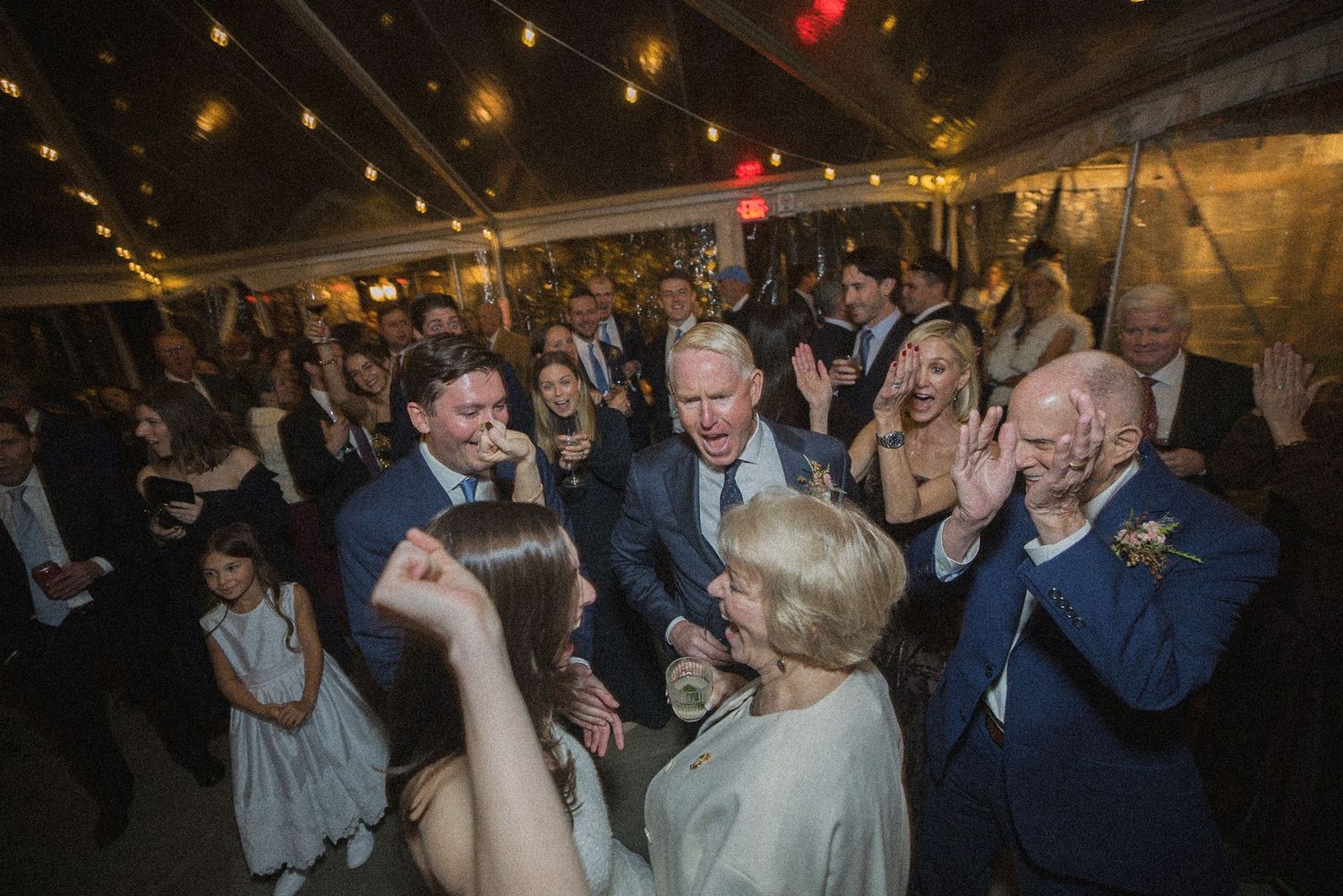 Guests in formal attire dance and clap together at an evening tent wedding reception under warm string lights.
