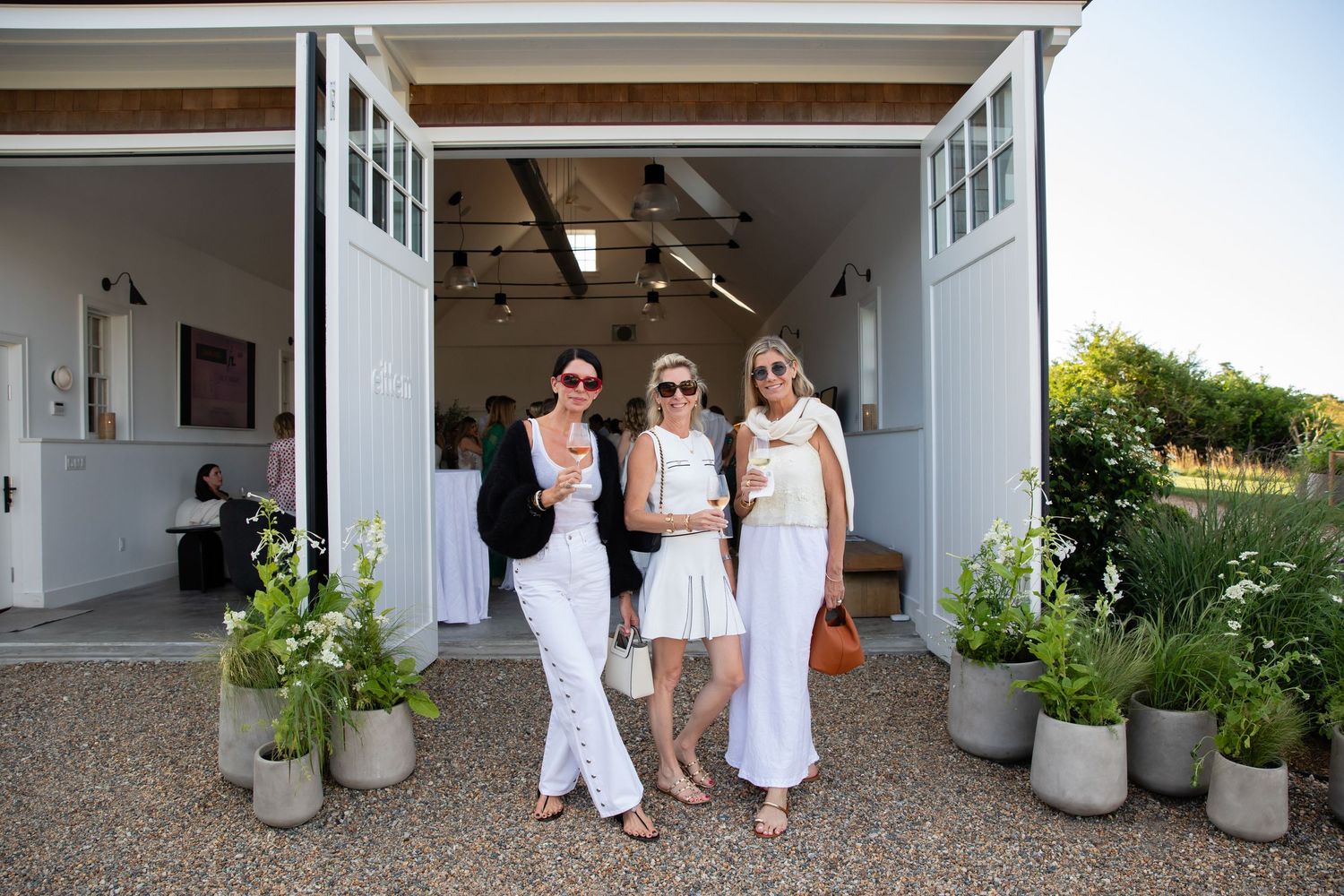 Three women in white outfits pose together outside a bright white winery tasting room surrounded by potted plants.