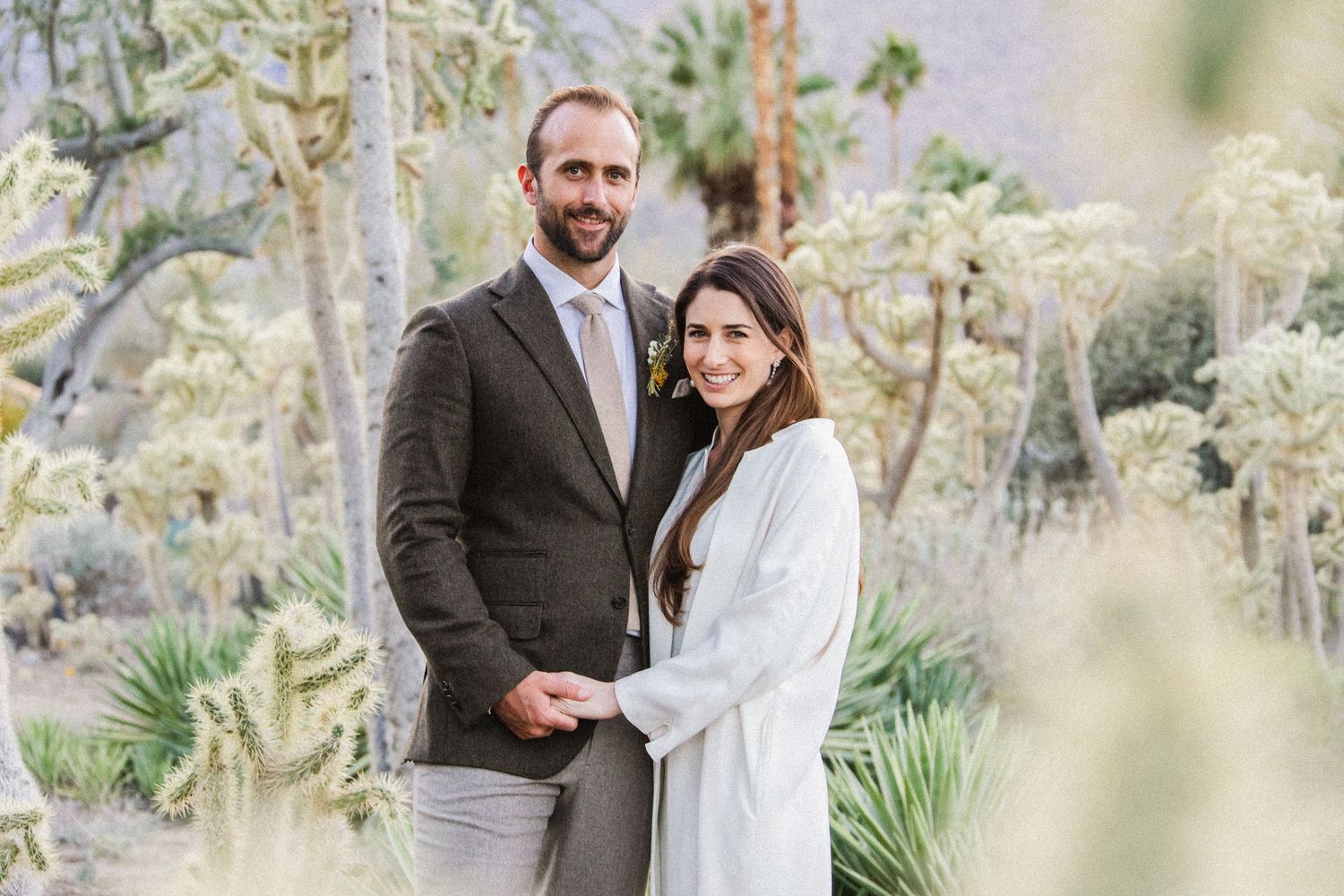 A couple stands together in a desert garden setting wearing wedding attire.