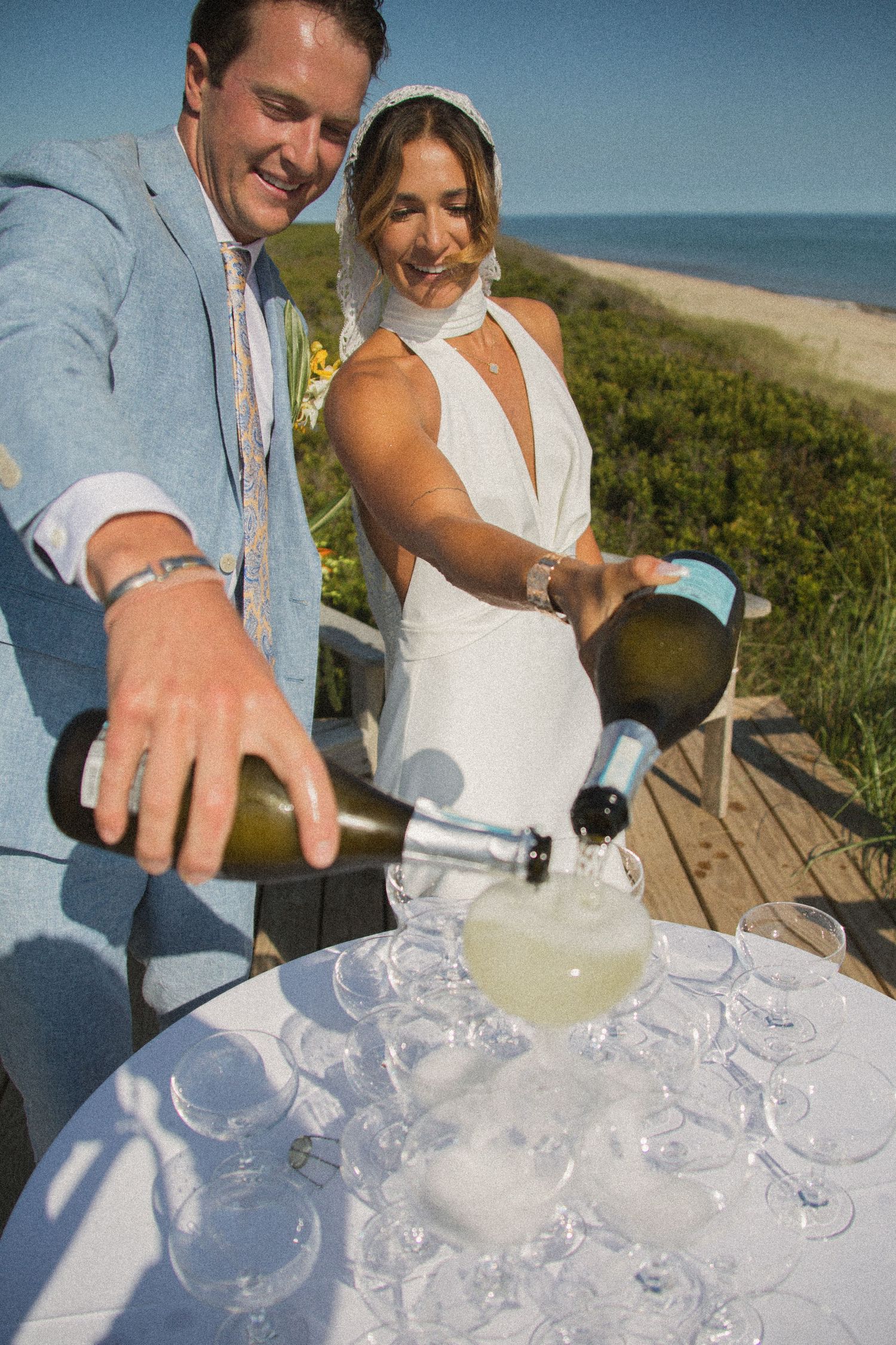 Pouring champagne into tower of glasses at outdoor beach celebration.