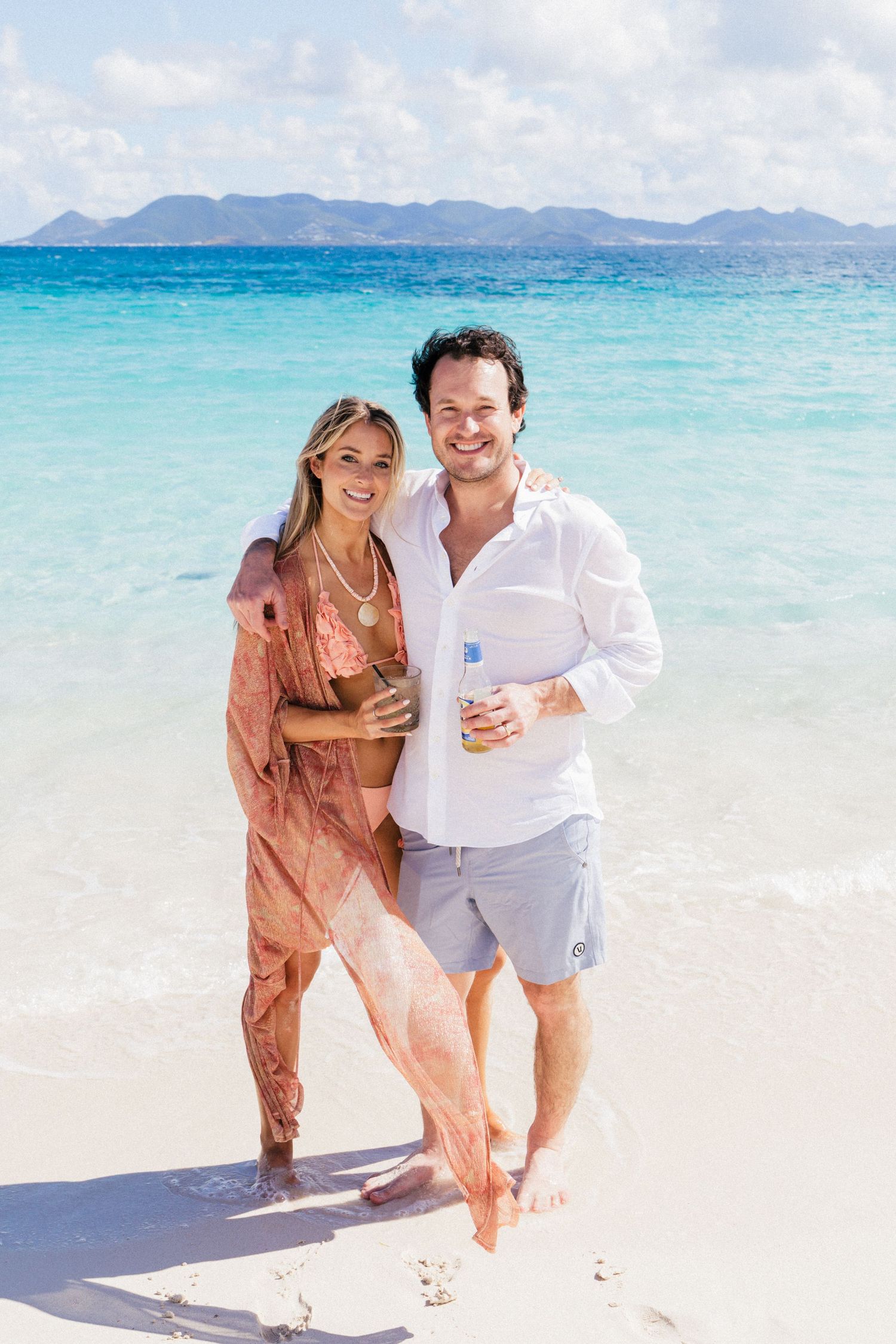 Couple embracing on tropical beach with turquoise waters and mountain landscape in background.
