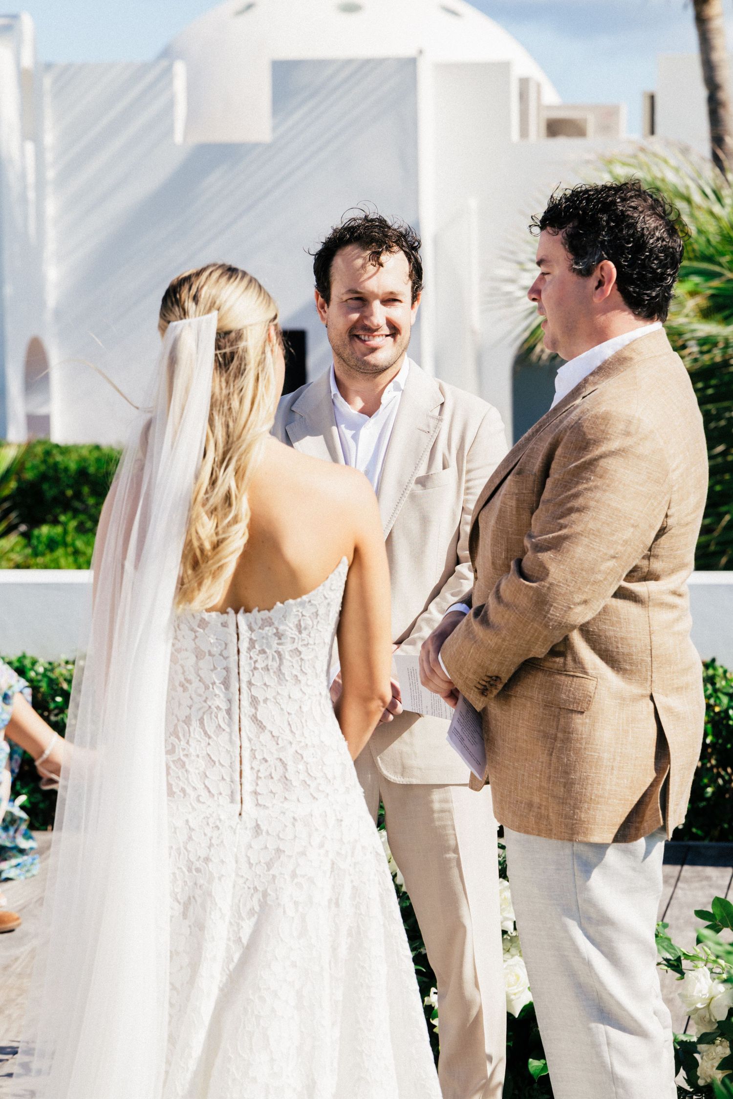 Wedding ceremony participants in light colored attire standing together on a sunny beach deck.