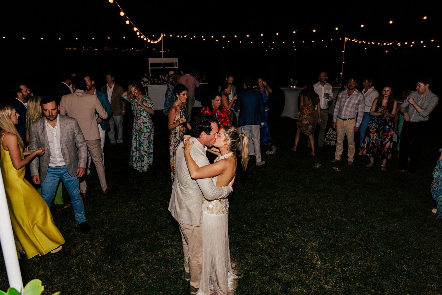 Wedding guests dance and celebrate on a grassy lawn under string lights at an outdoor evening reception.