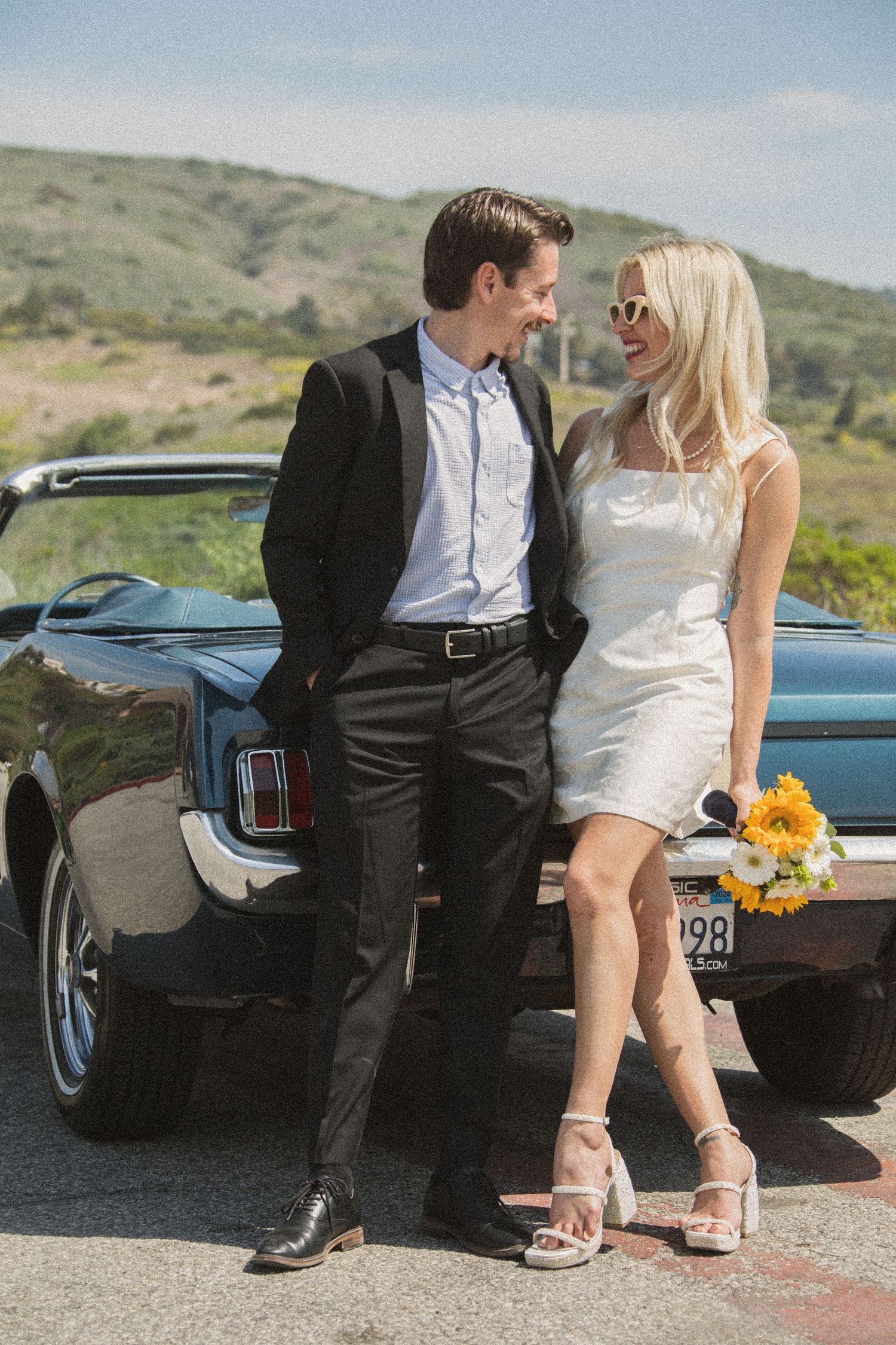 Romantic photoshoot featuring a couple sitting on a vintage Mustang with yellow flowers.
