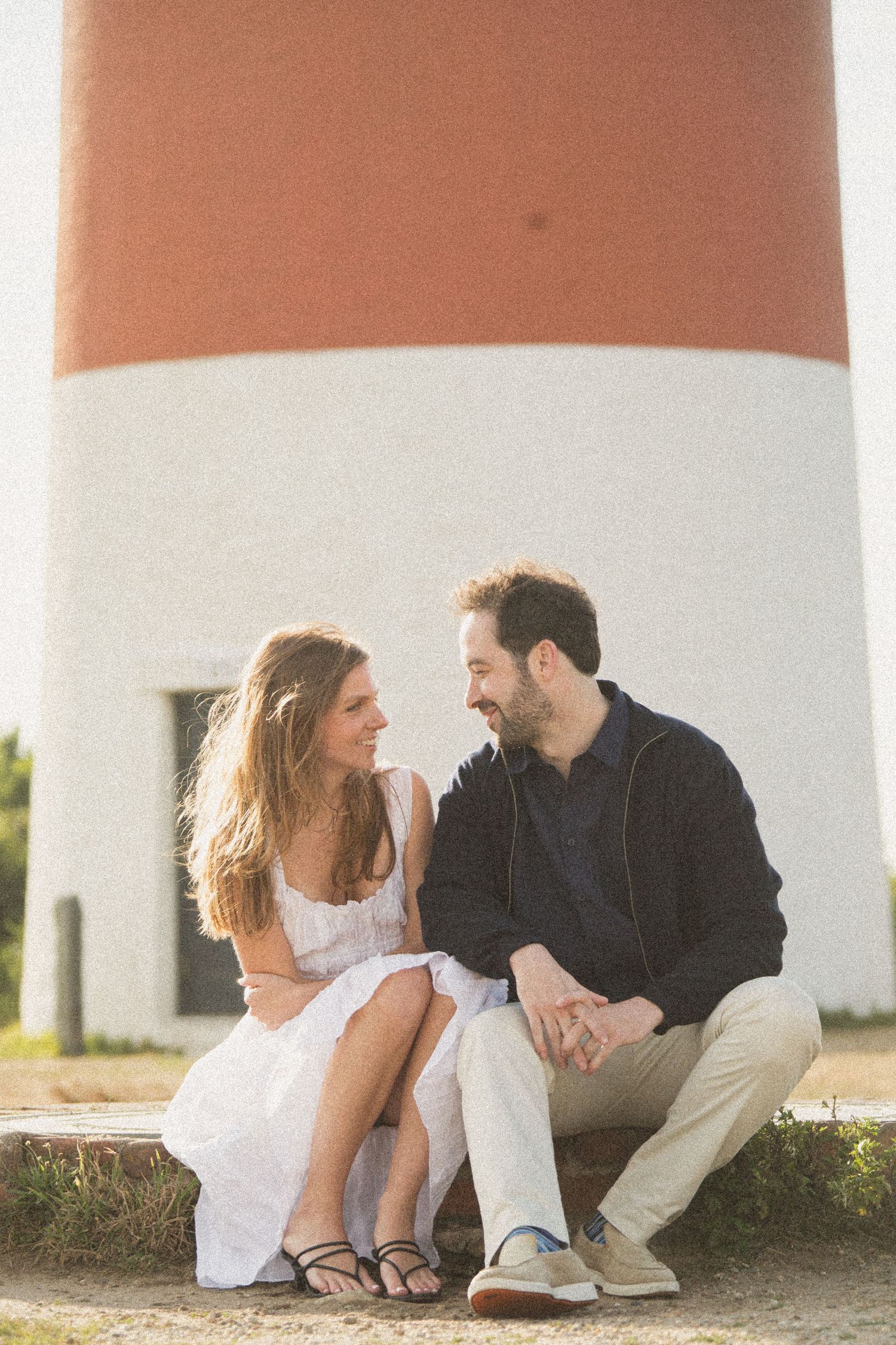 Sunset lighthouse engagement session with couple sitting on ground.