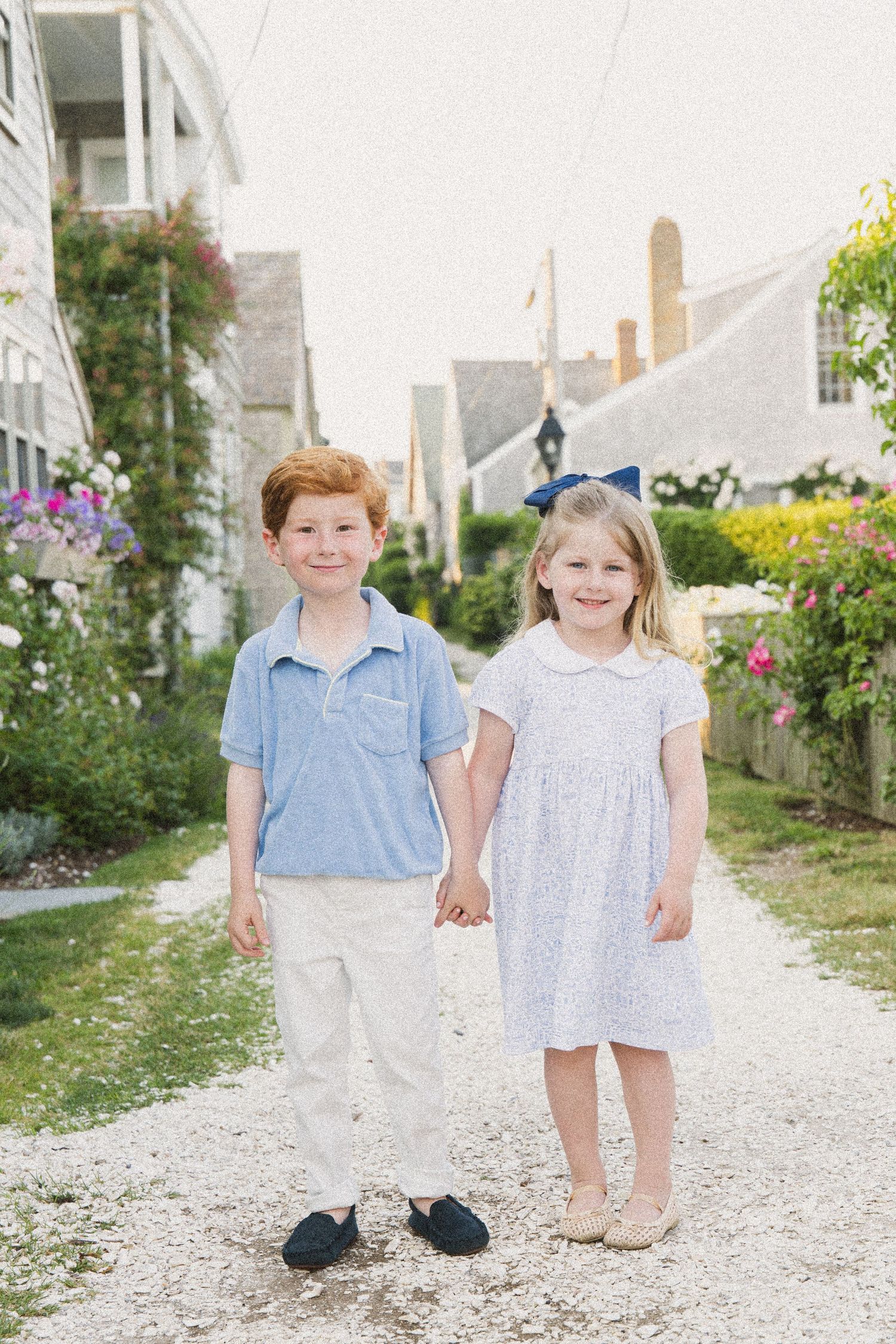 Two young children holding hands on a gravel path, boy in blue polo and girl in floral dress with blue bow.