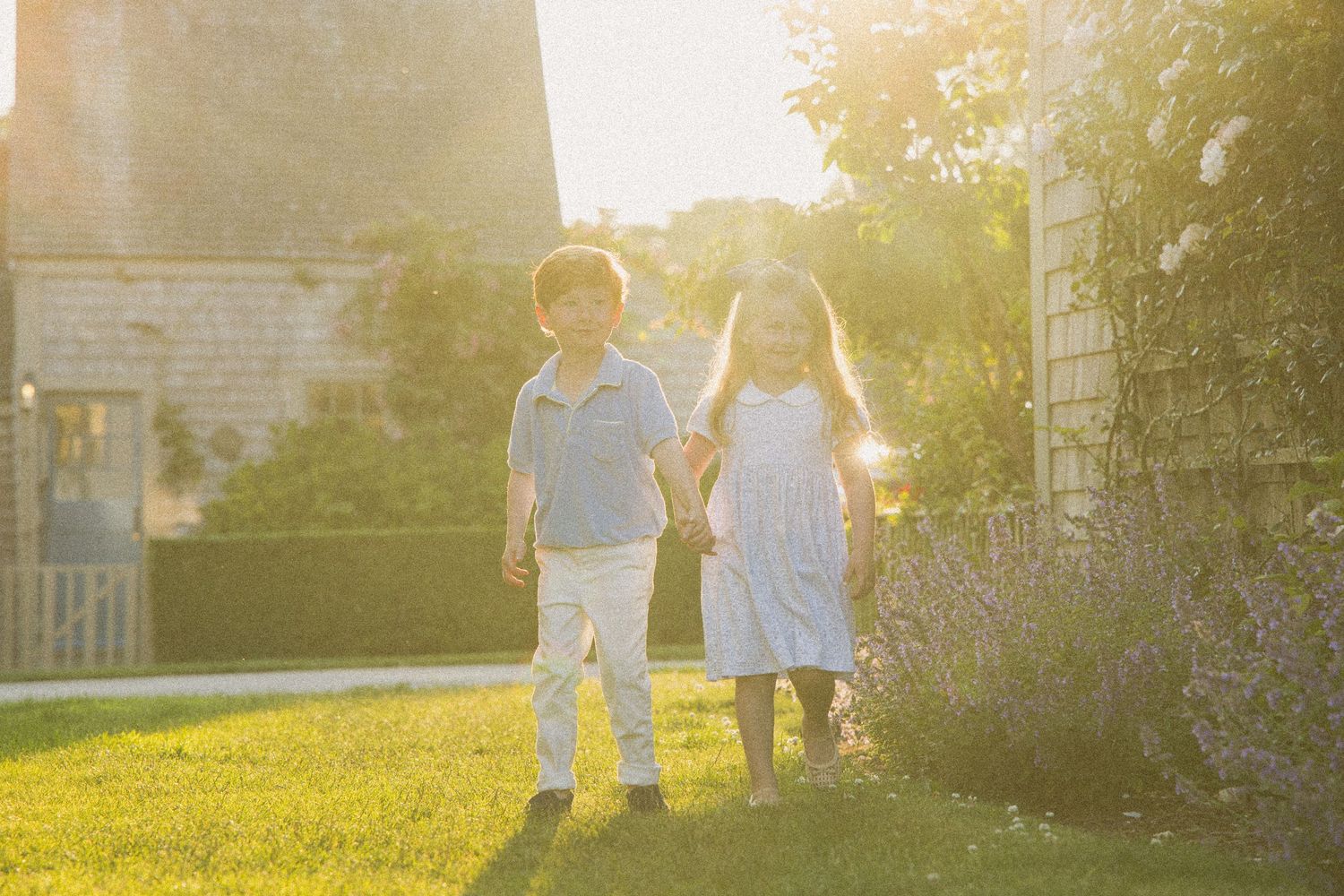 Two children holding hands in a sunlit garden, boy in blue polo and girl in white dress, golden hour light.