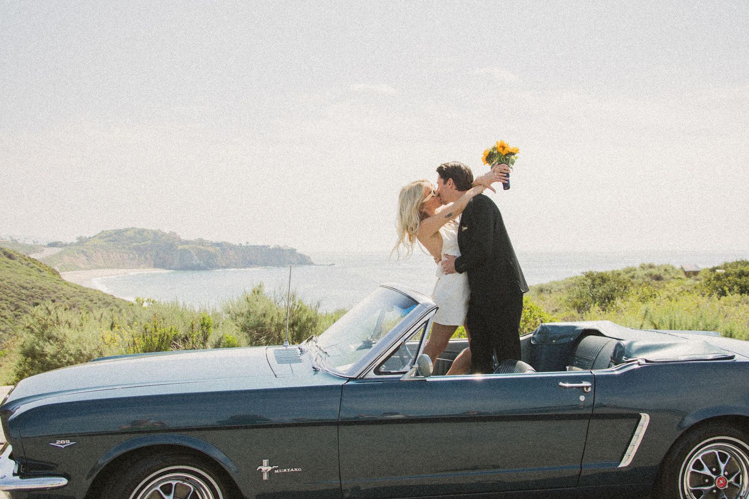Newlyweds kissing on a vintage blue Ford Mustang convertible with ocean views and yellow bouquet.