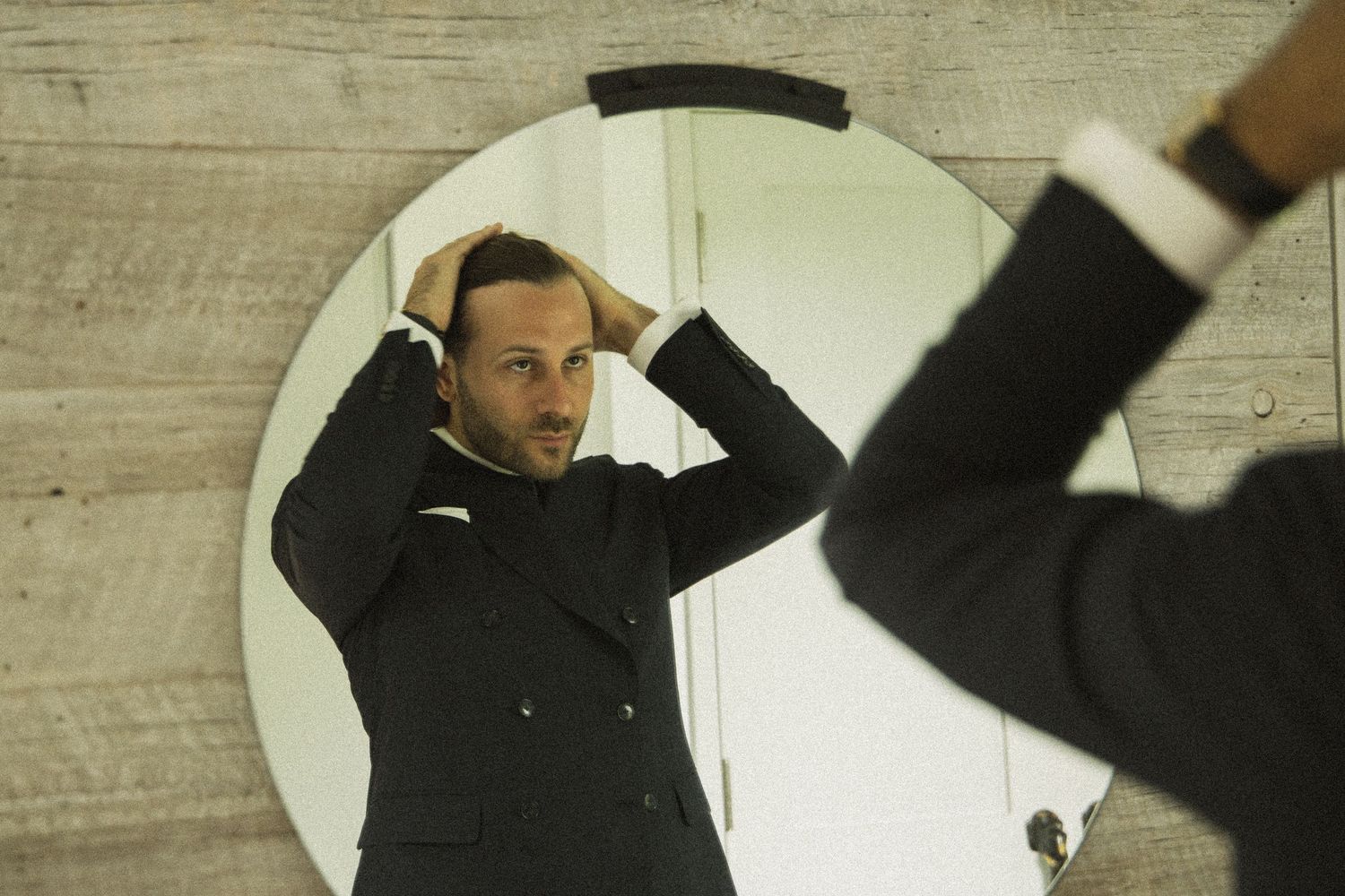 Man in dark suit fixing his hair while looking at his reflection in a round wall mirror.