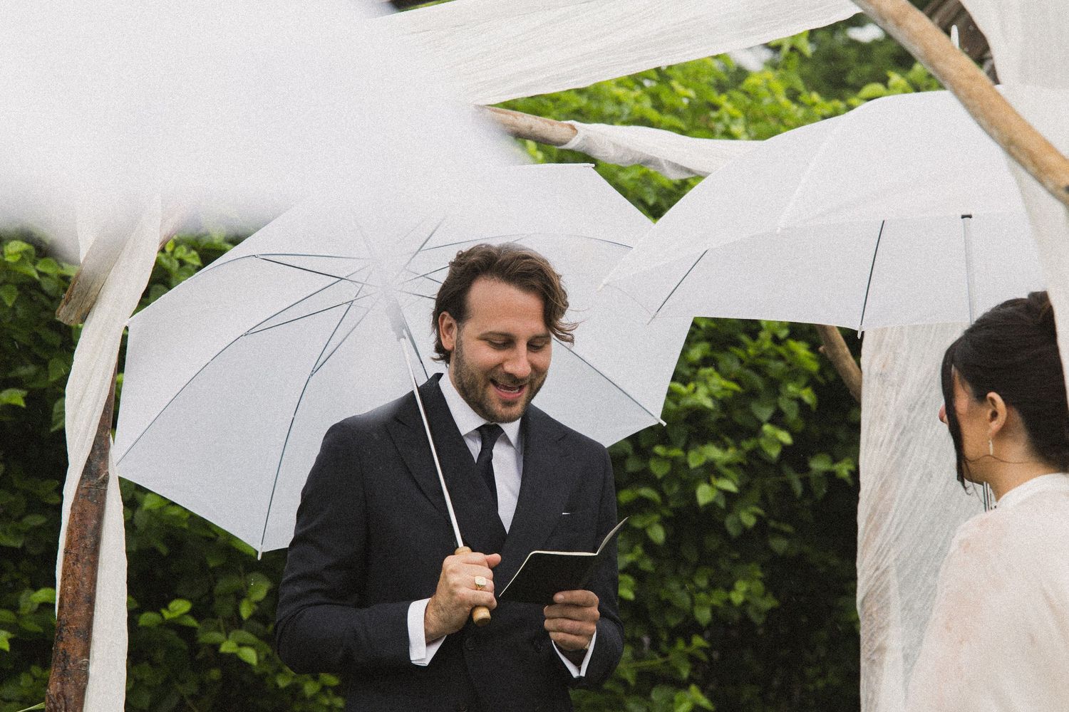 Man in black suit holding white umbrella and reading at outdoor wedding ceremony with white draping.
