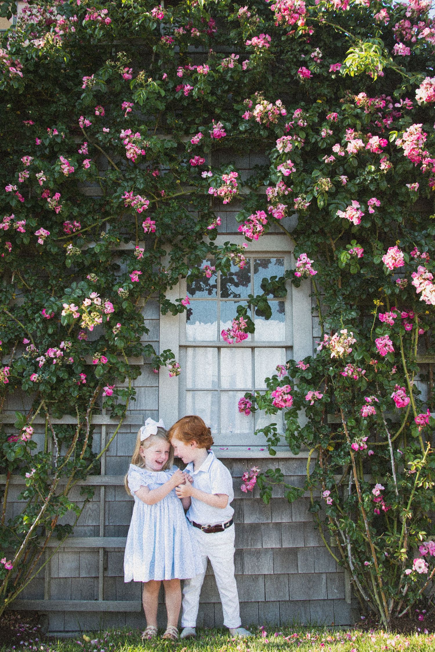 Children posing by climbing rose bushes against a gray shingled house.