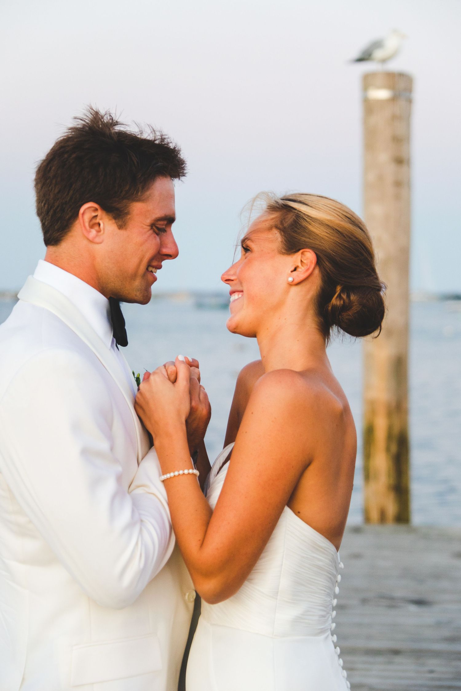 A couple shares a joyful moment on a waterfront dock in white wedding attire.