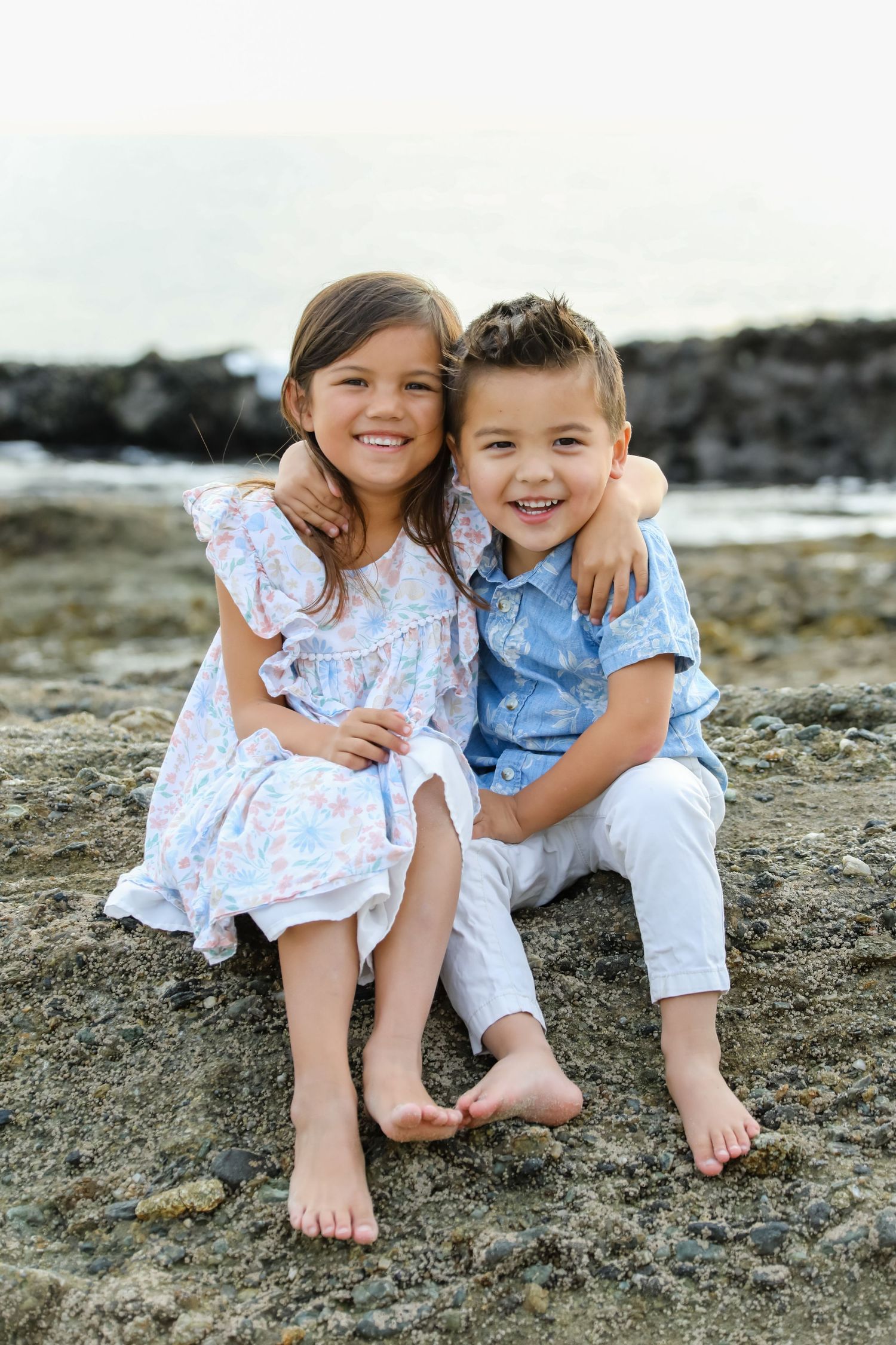 Two children in coordinating blue and white outfits sit together on coastal rocks by the ocean.