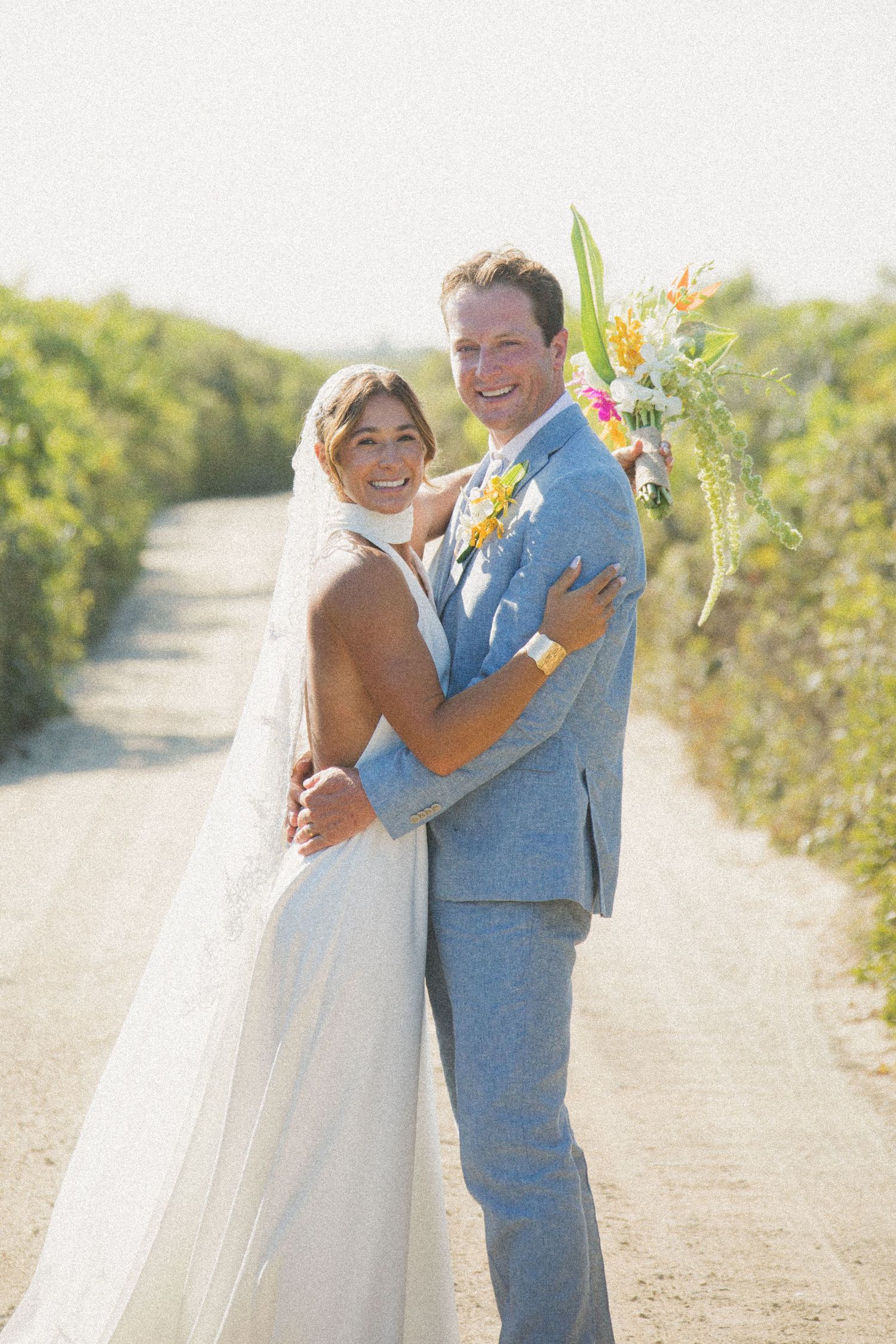Happy couple embraces on a sunny path during their beach wedding photoshoot.