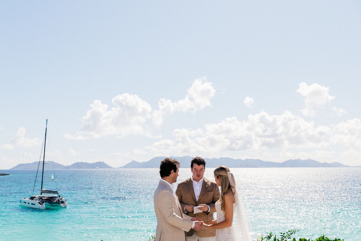 Destination wedding scene with mountains and sailboats visible across the tranquil ocean waters.