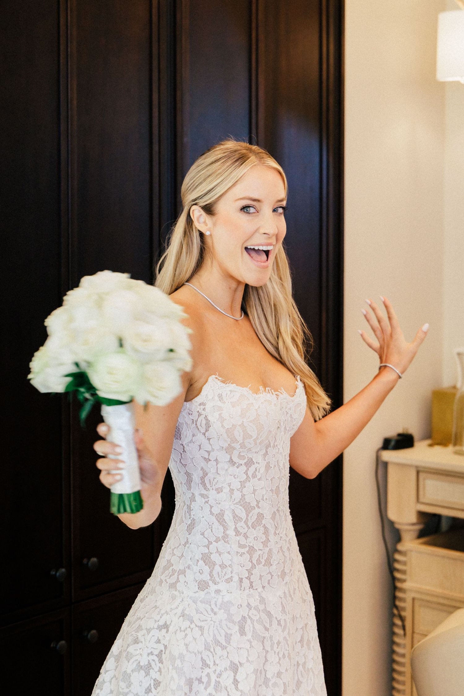 Bride in lace gown holds white bouquet while posing against dark doors in elegant interior.