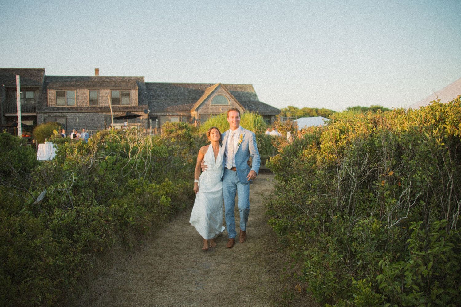 Wedding party walks along a beach path with houses visible in the background.