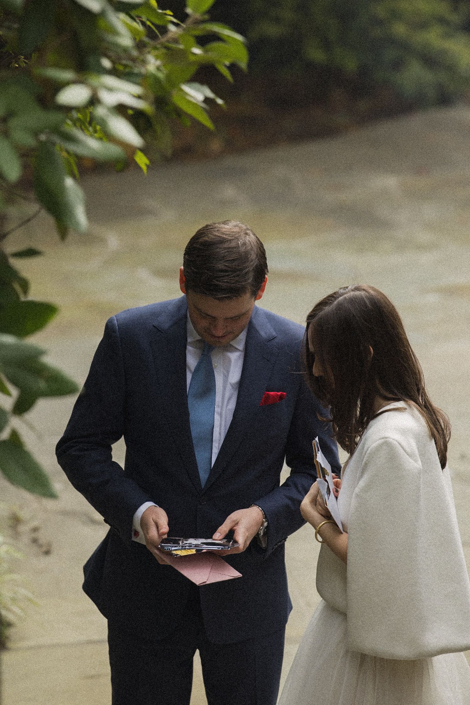 Groom in a navy suit and blue tie reads a card beside a bride in a white wrap while standing near a stream.