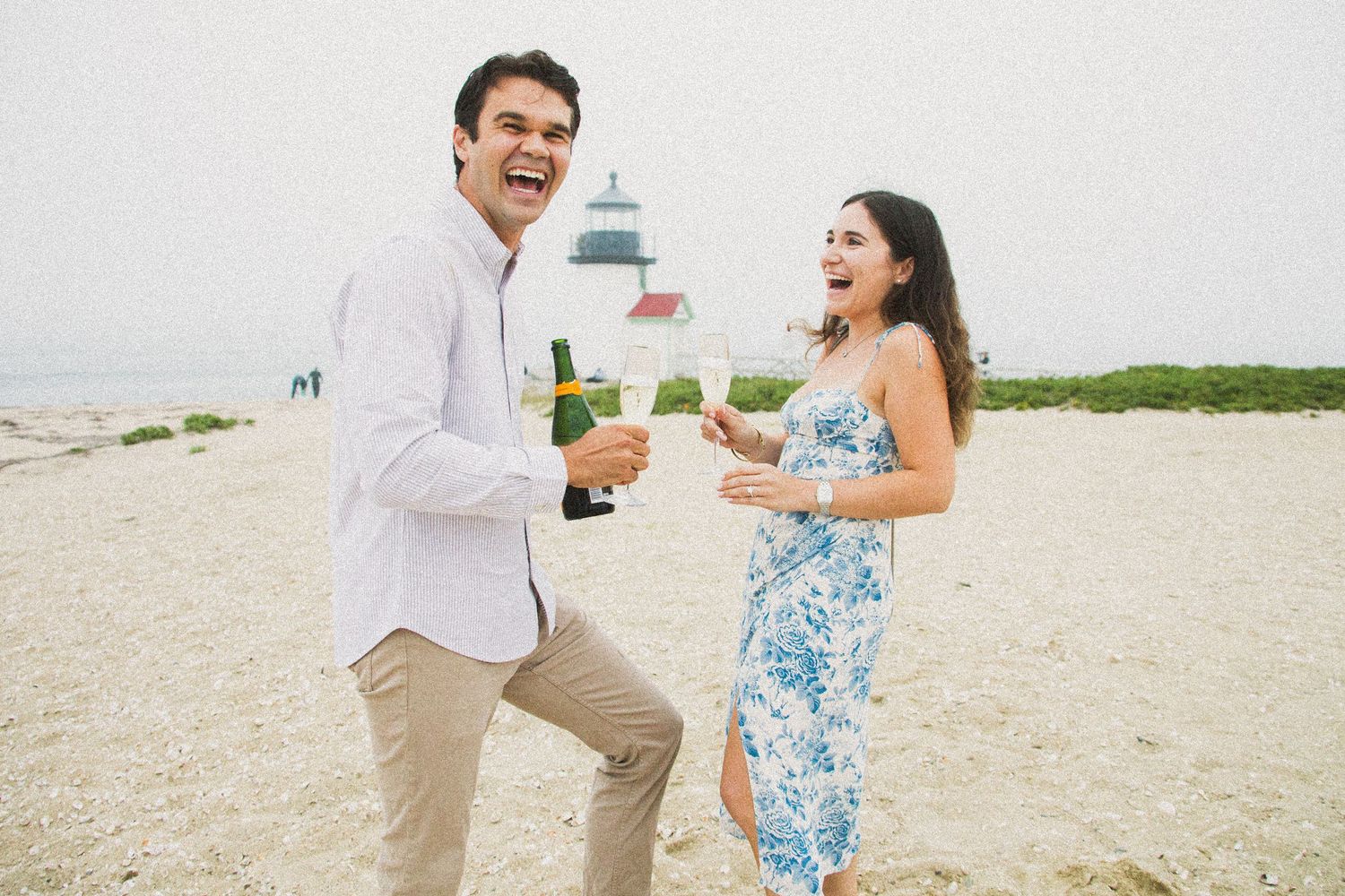 Couple laughing on beach holding champagne bottles near lighthouse on overcast day.