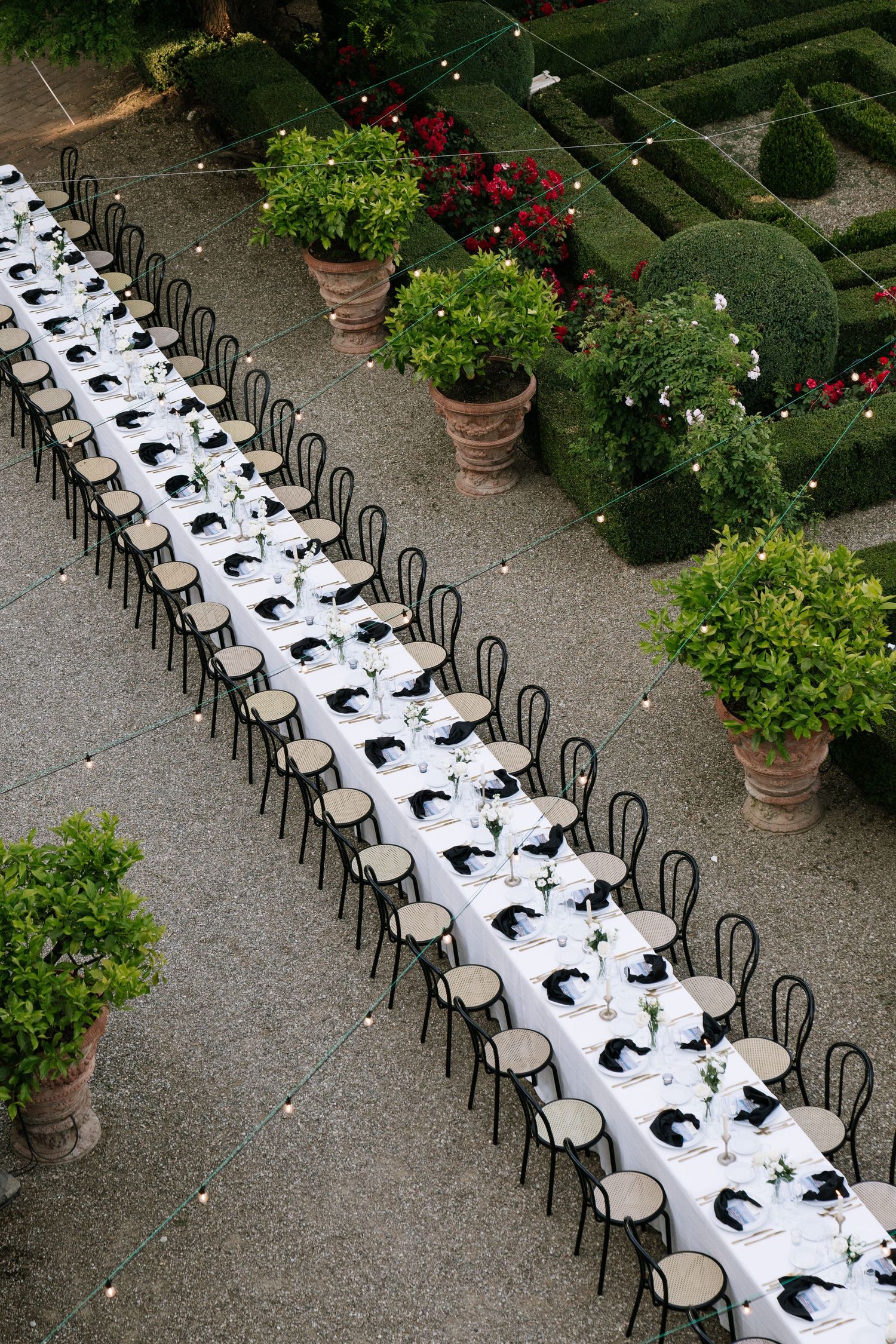Aerial view of elegant outdoor wedding reception with long white-draped tables, black chairs, and lush garden greenery.
