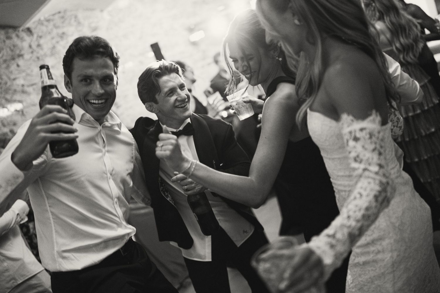 Black and white photo of wedding guests dancing and celebrating joyfully, bride in lace dress visible.