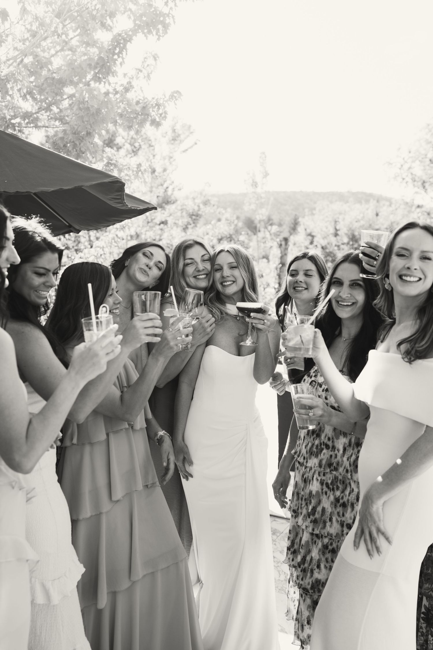 Black and white photo of a bride and bridesmaids toasting with drinks outdoors at a wedding celebration.