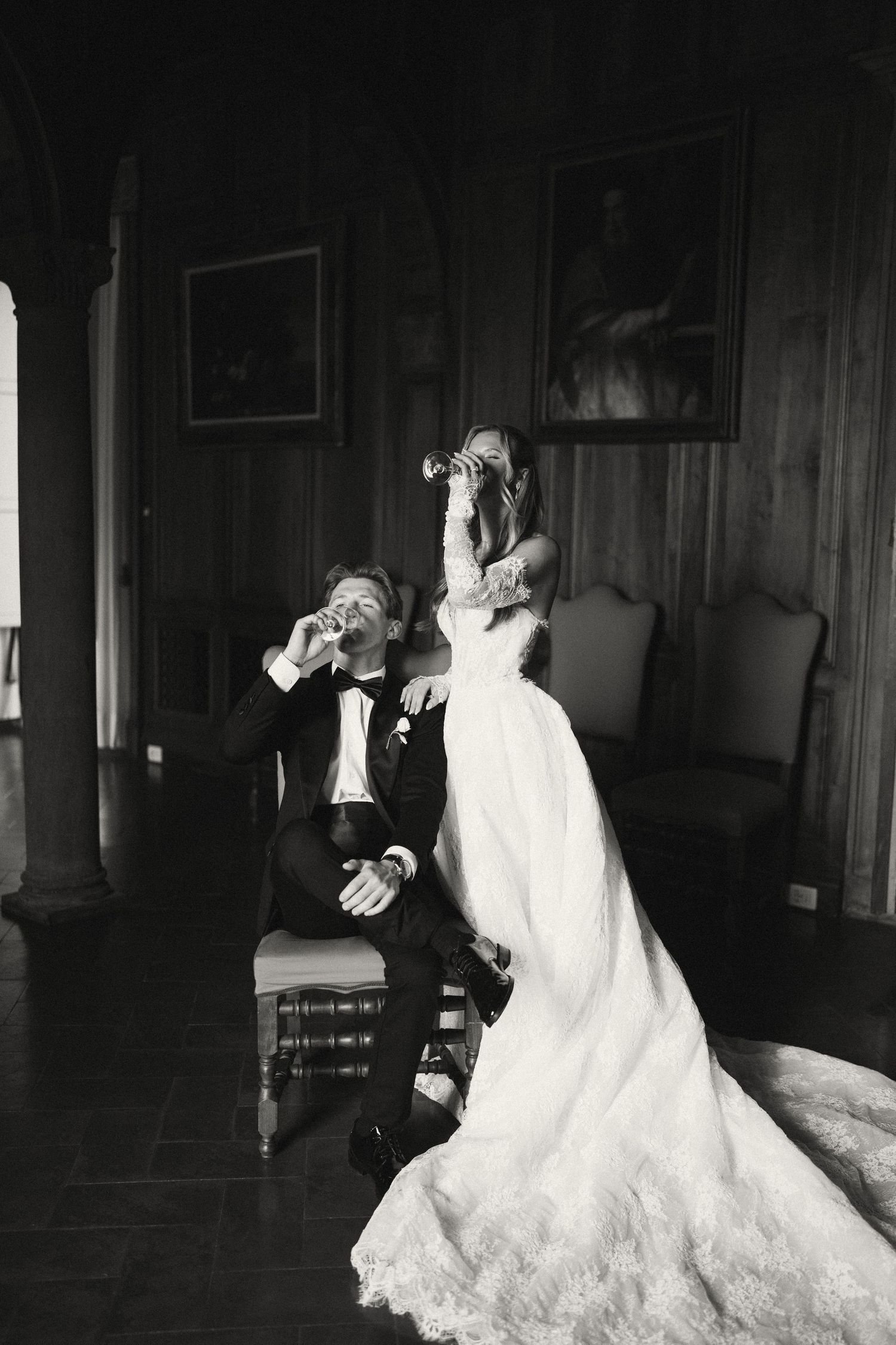 Black and white wedding photo of bride and groom in elegant room, groom drinking as bride photographs him.