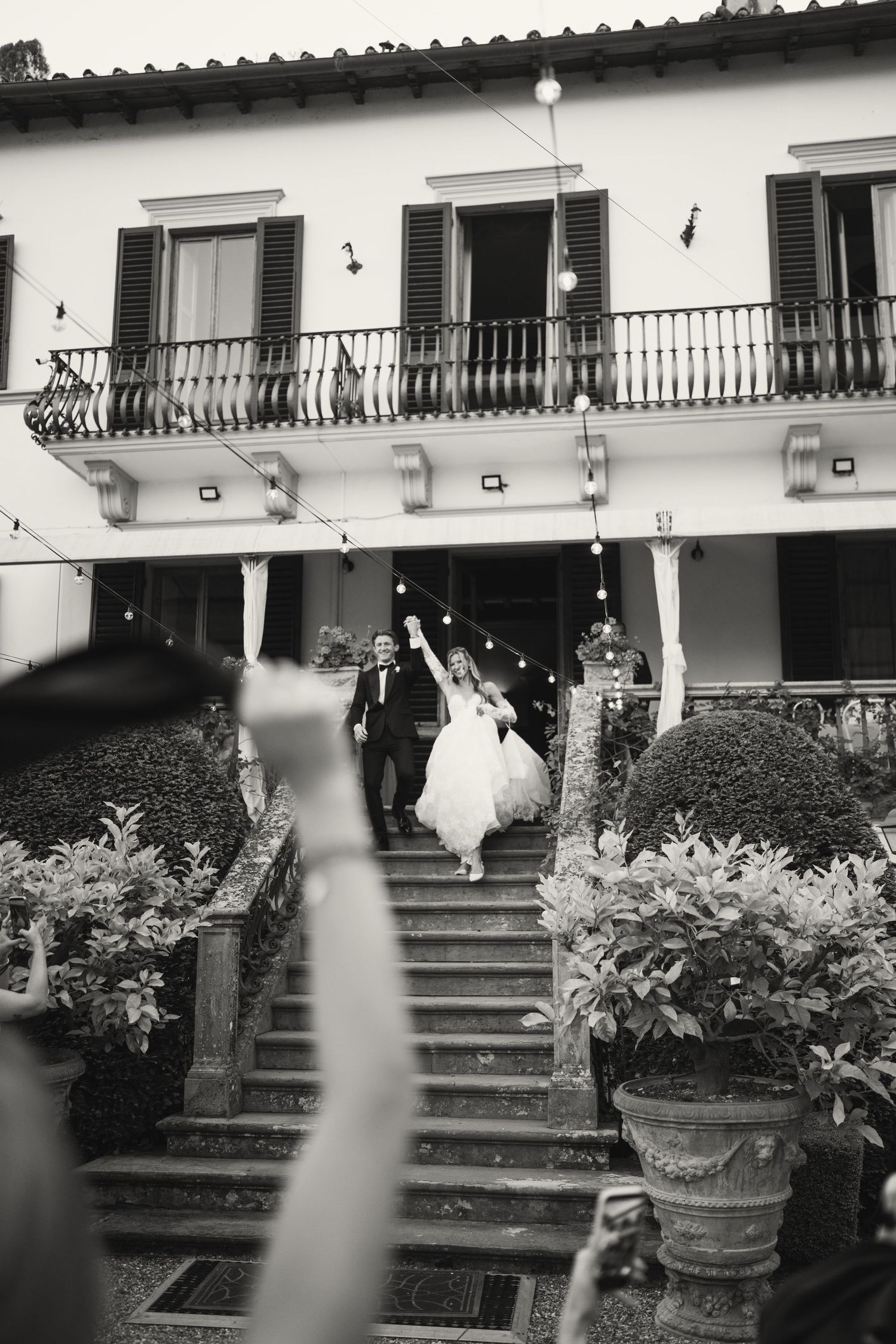 Black and white photo of a bride and groom descending grand steps of an Italian villa while guests celebrate below.