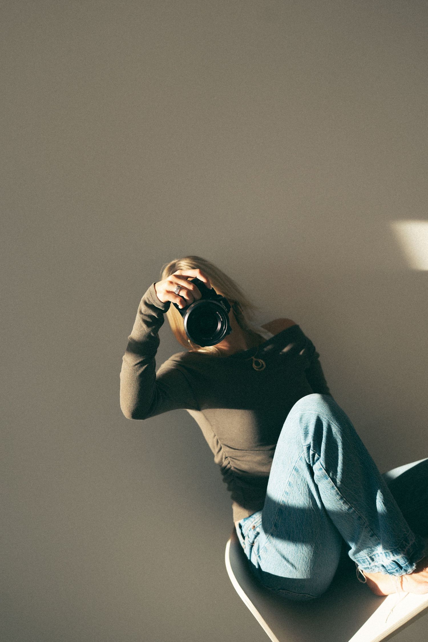 Person holding camera while sitting, wearing gray top and light jeans in minimalist setting.