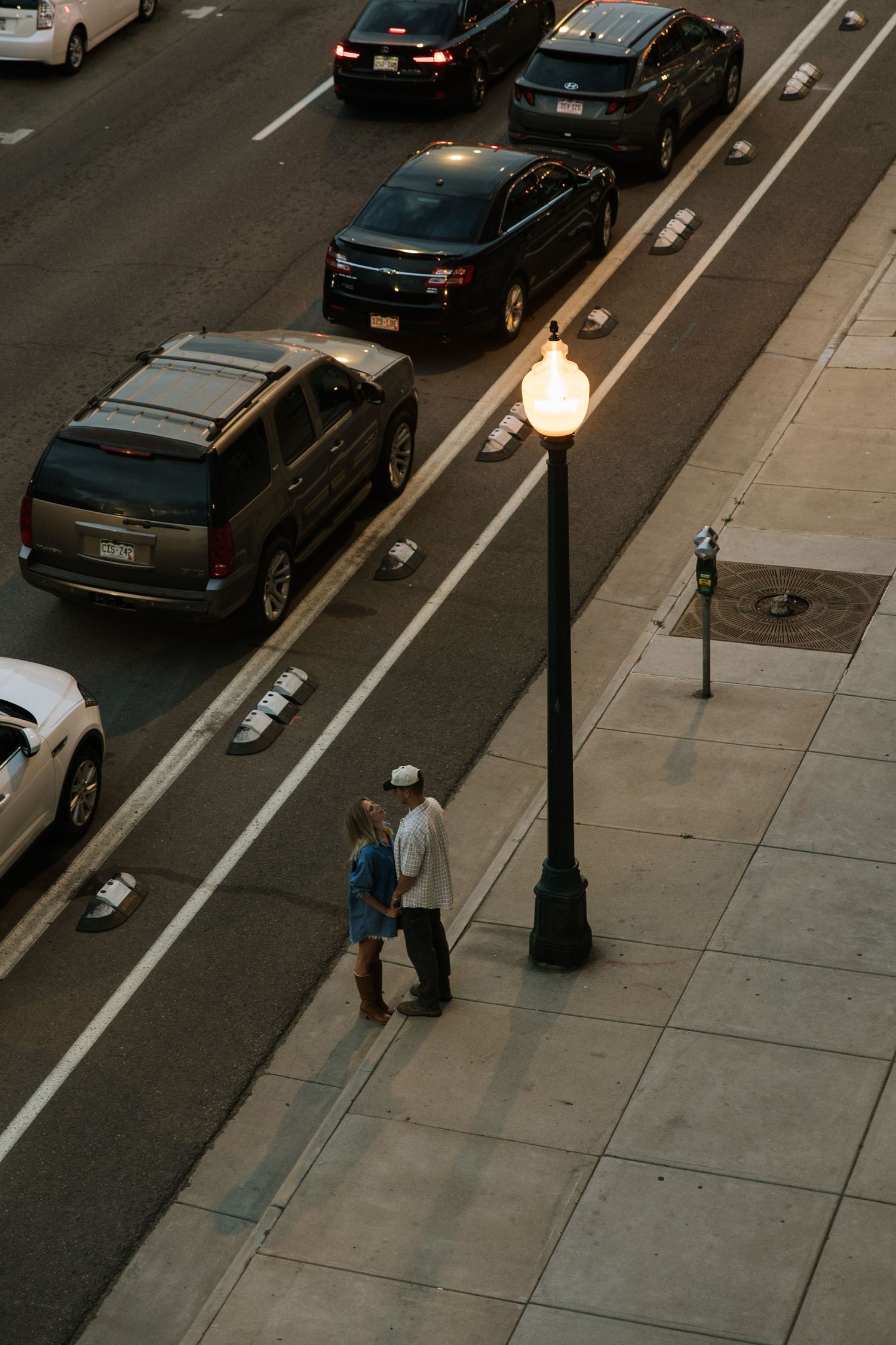People walking on a city sidewalk next to parked cars and street lamps at dusk.