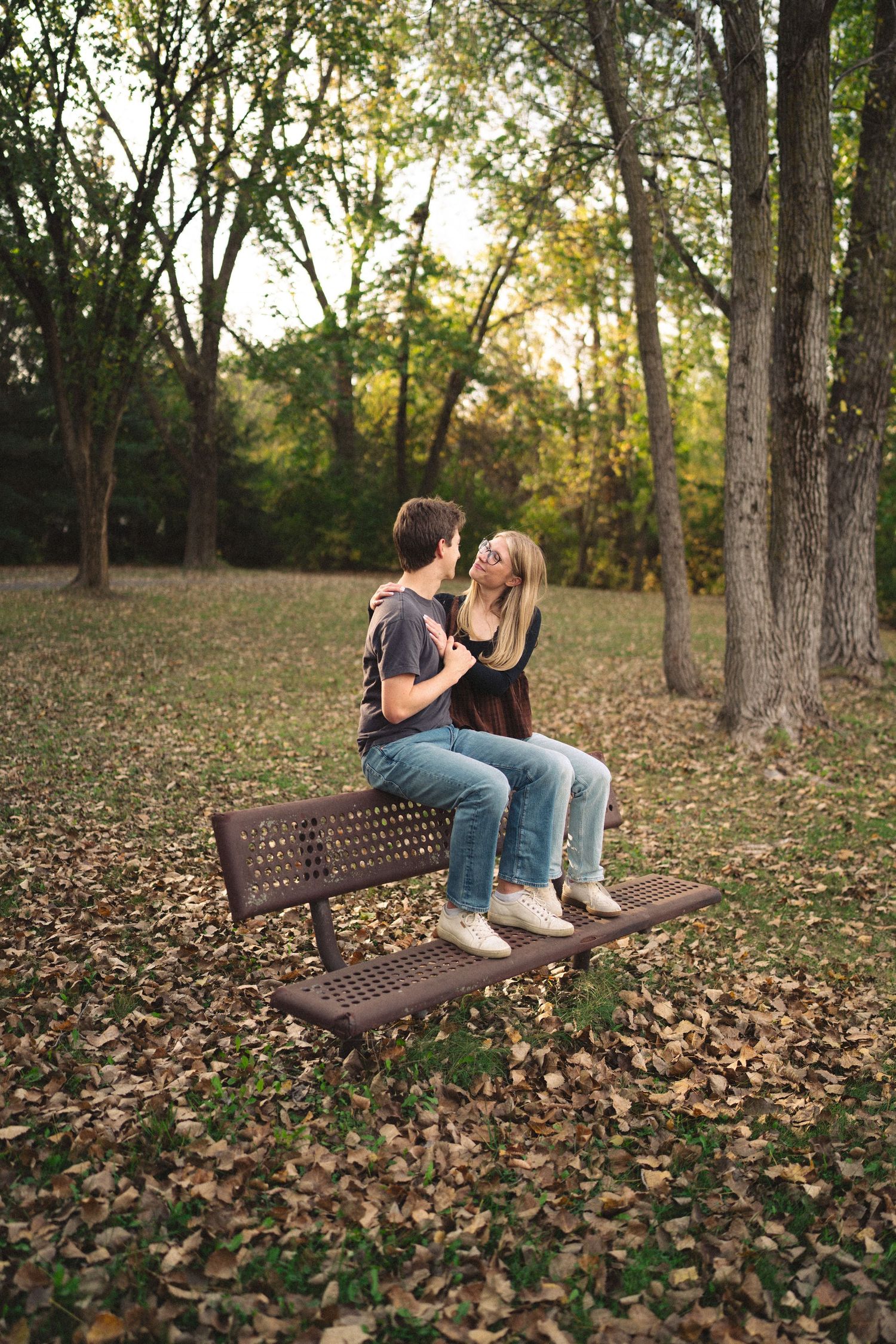 A couple sharing a tender moment while sitting on a park bench surrounded by fallen autumn leaves.
