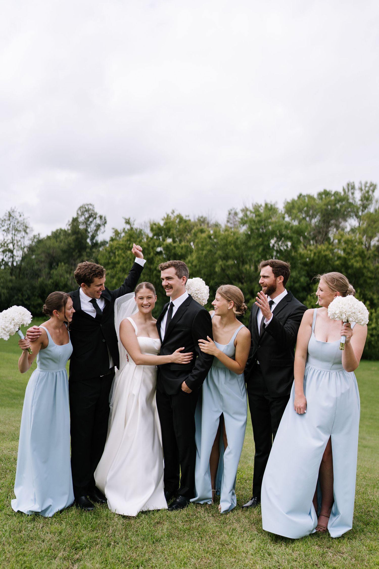 Wedding party in light blue bridesmaid dresses and black suits posing together on grass.