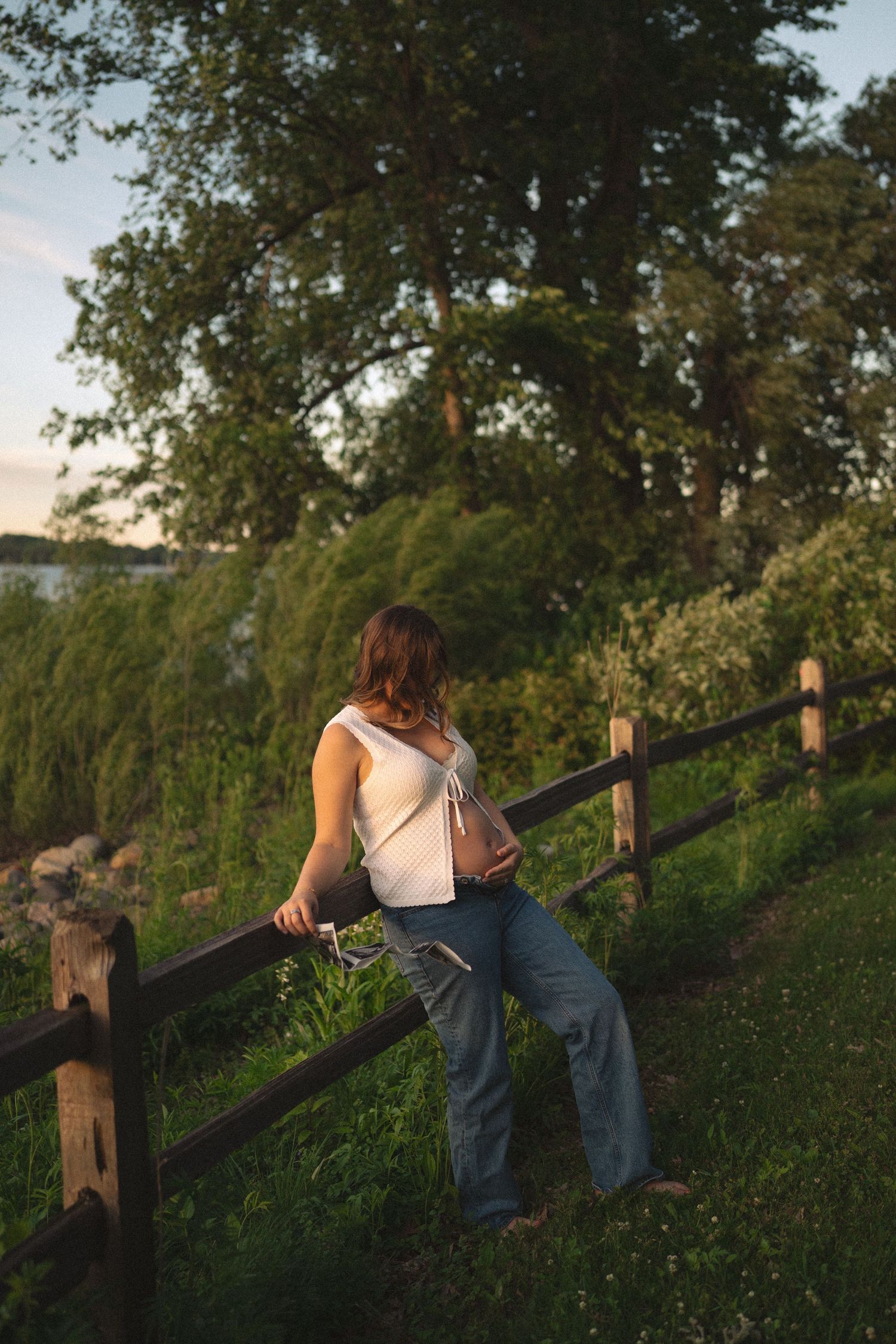 Woman in jeans and white top leaning on wooden fence surrounded by lush green trees at golden hour sunset.