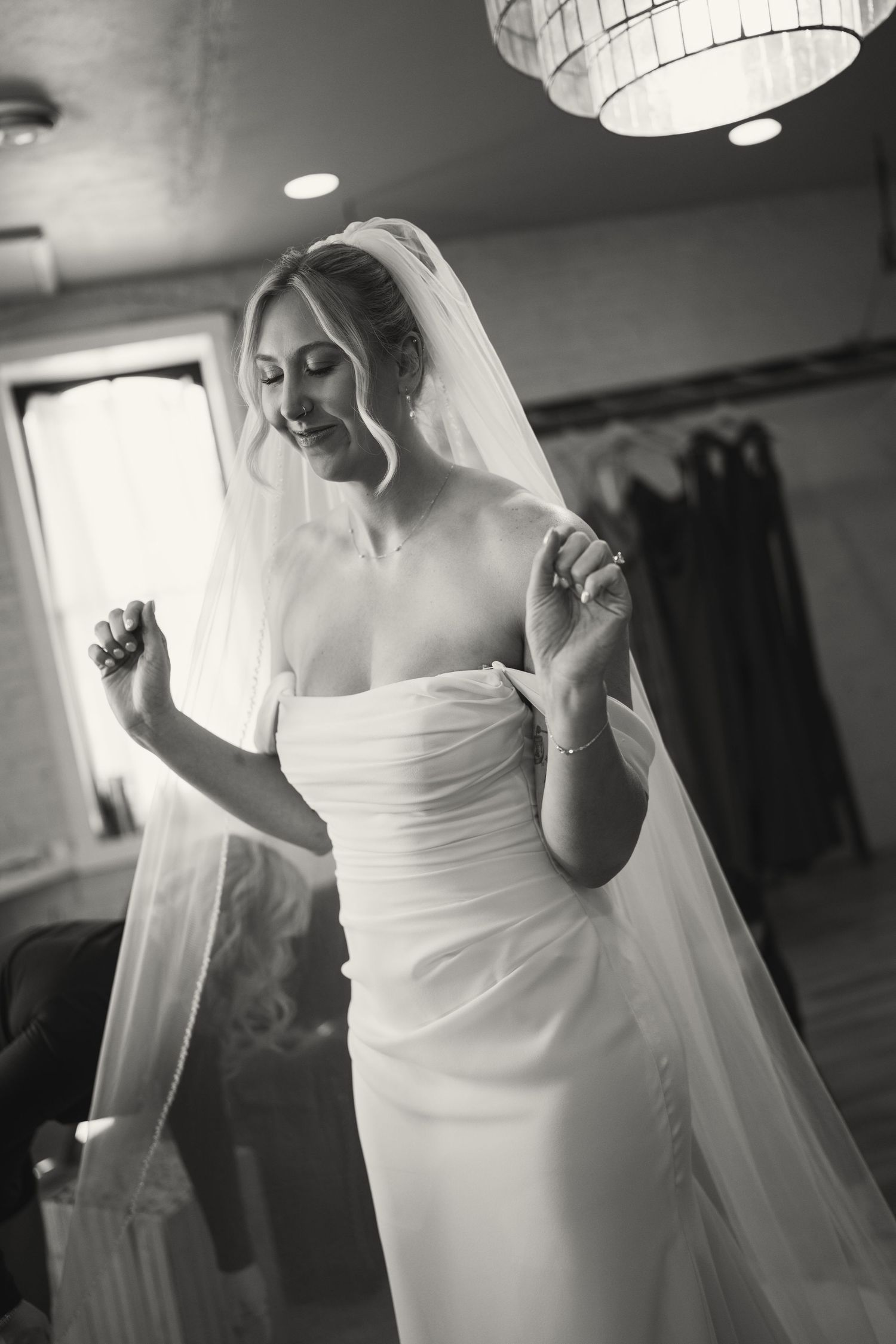 Bride in white dress adjusts veil under soft lighting fixture.