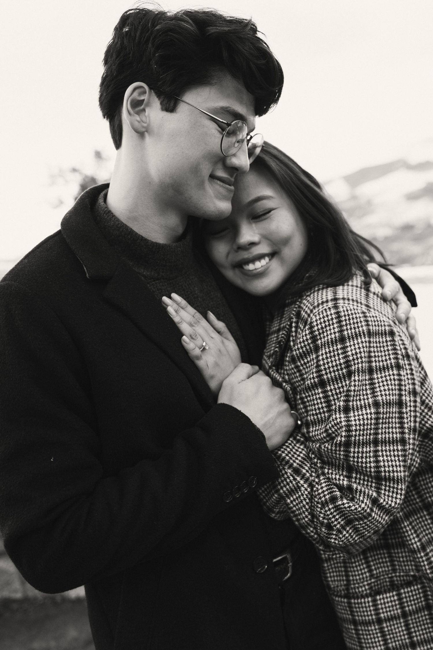 Black and white photo of a smiling couple embracing outdoors, woman in plaid coat hugging man in glasses and dark jacket.