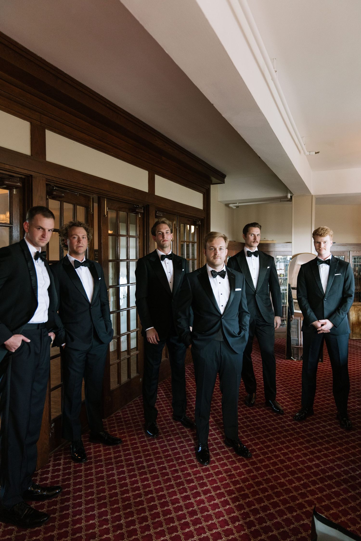 Groomsmen in classic black tuxedos pose in an elegant hallway.