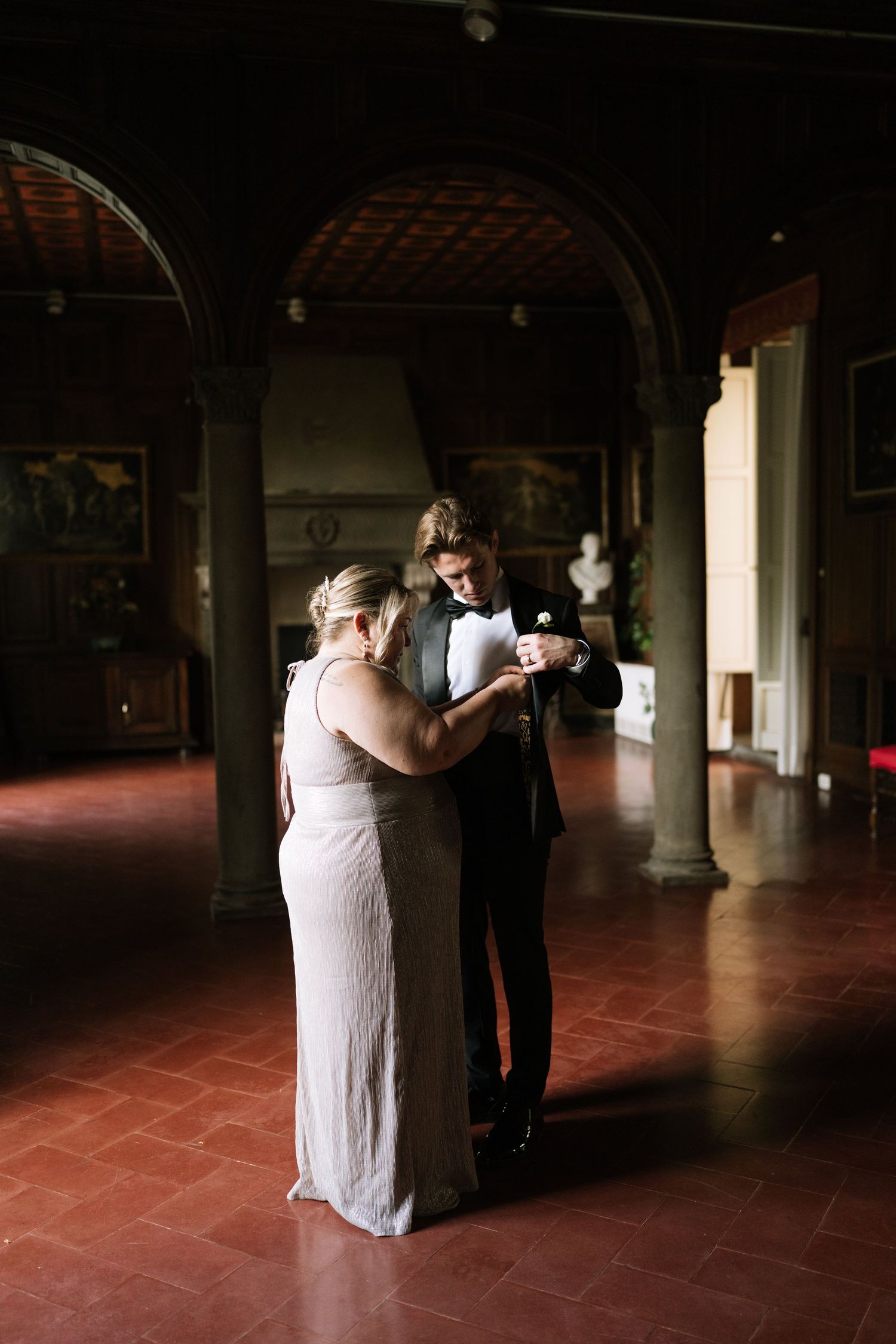 Groom in black tuxedo shares a tender moment with a woman in a elegant hall with arched columns.