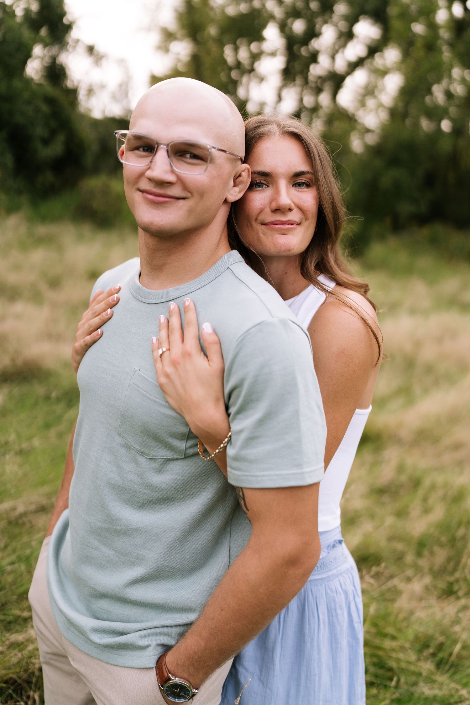 A couple poses together in casual attire against a backdrop of trees and grass.