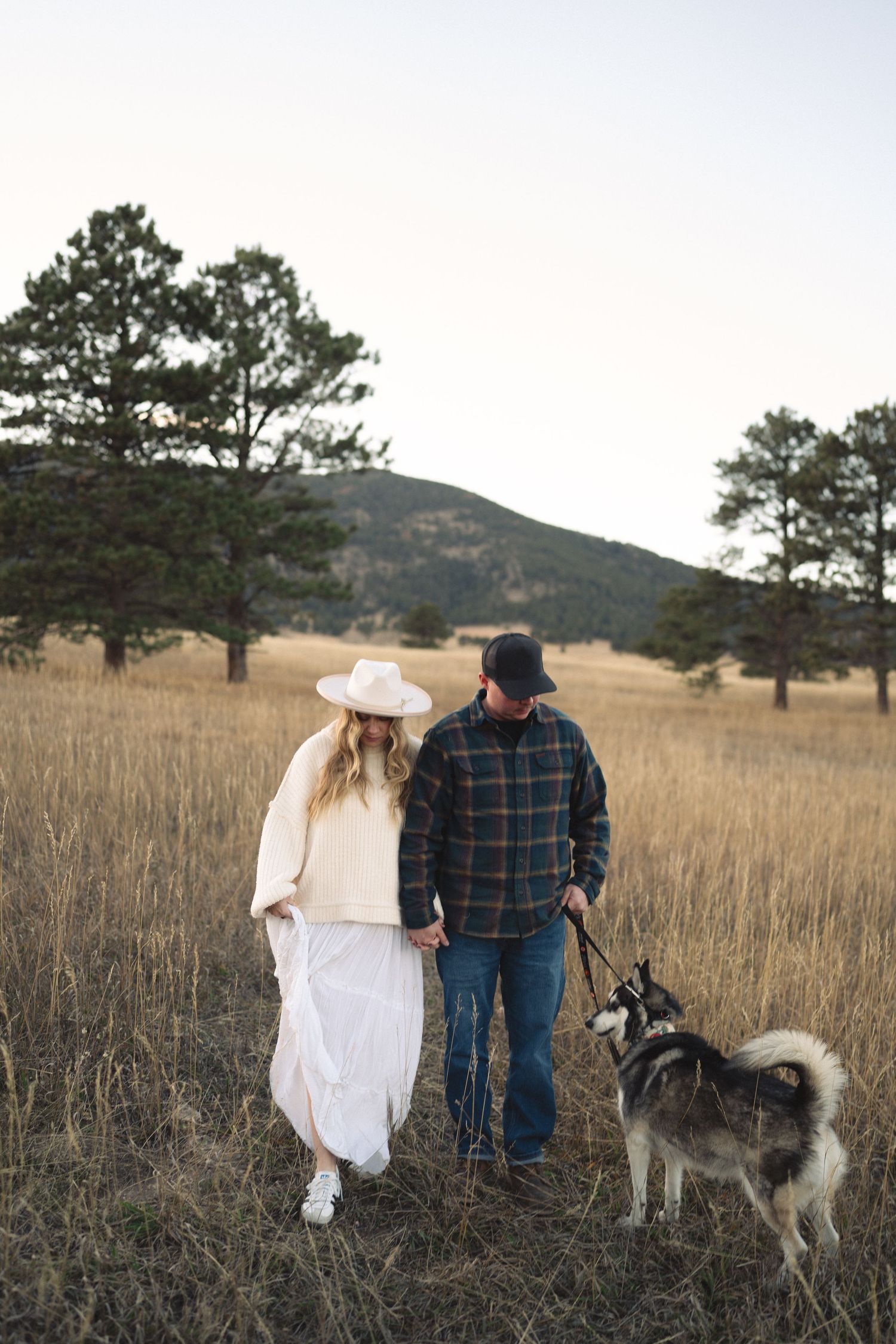 Couple walks with husky dog through mountain meadow at sunset.
