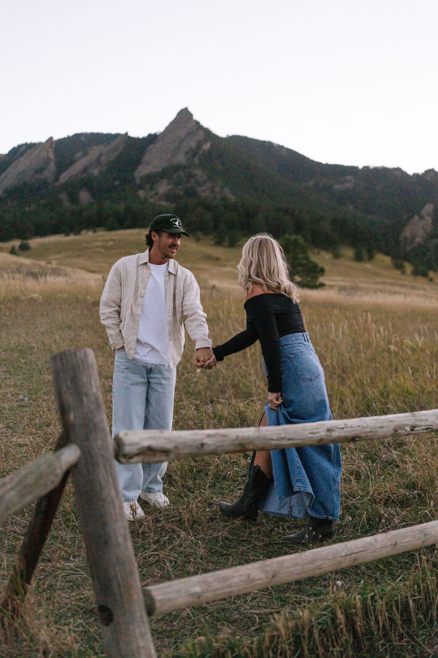 A couple sharing a moment while leaning against a wooden fence in a mountain landscape.