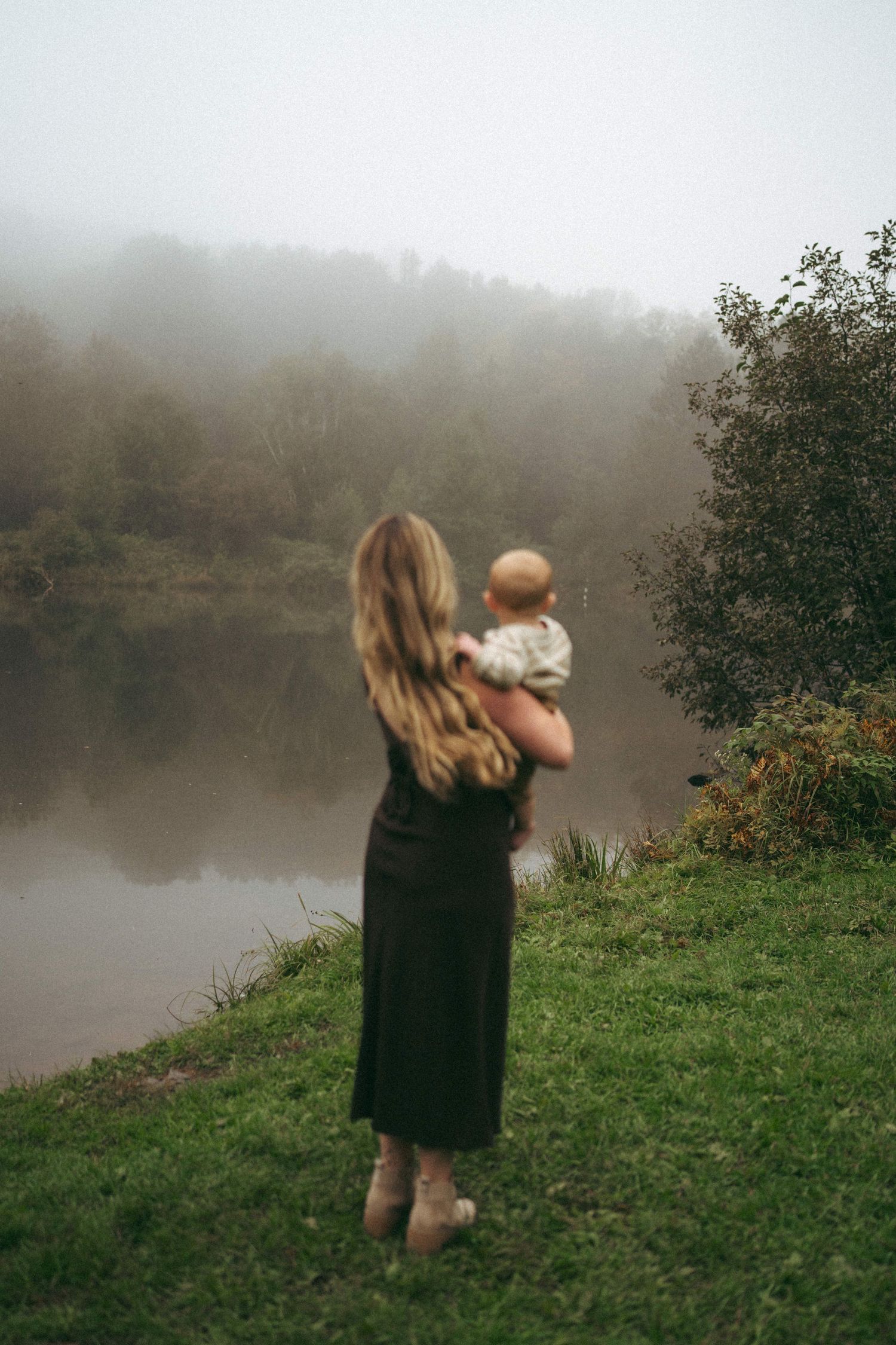 Woman holding baby while looking out over misty lake on autumn morning.