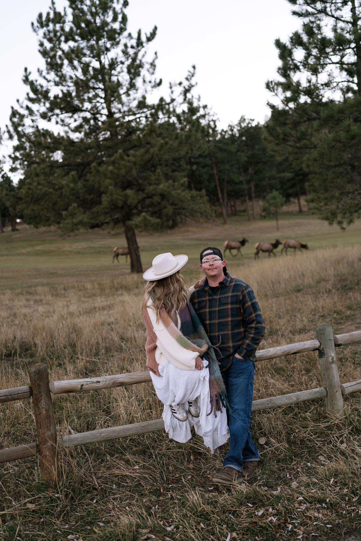 A couple stands by a wooden fence while elk graze in the background meadow.