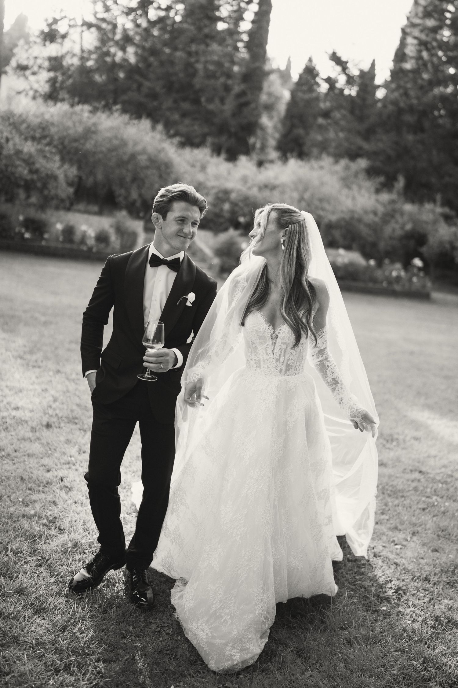 Black and white photo of a bride and groom walking in a garden, bride in lace gown with veil, groom in tuxedo.