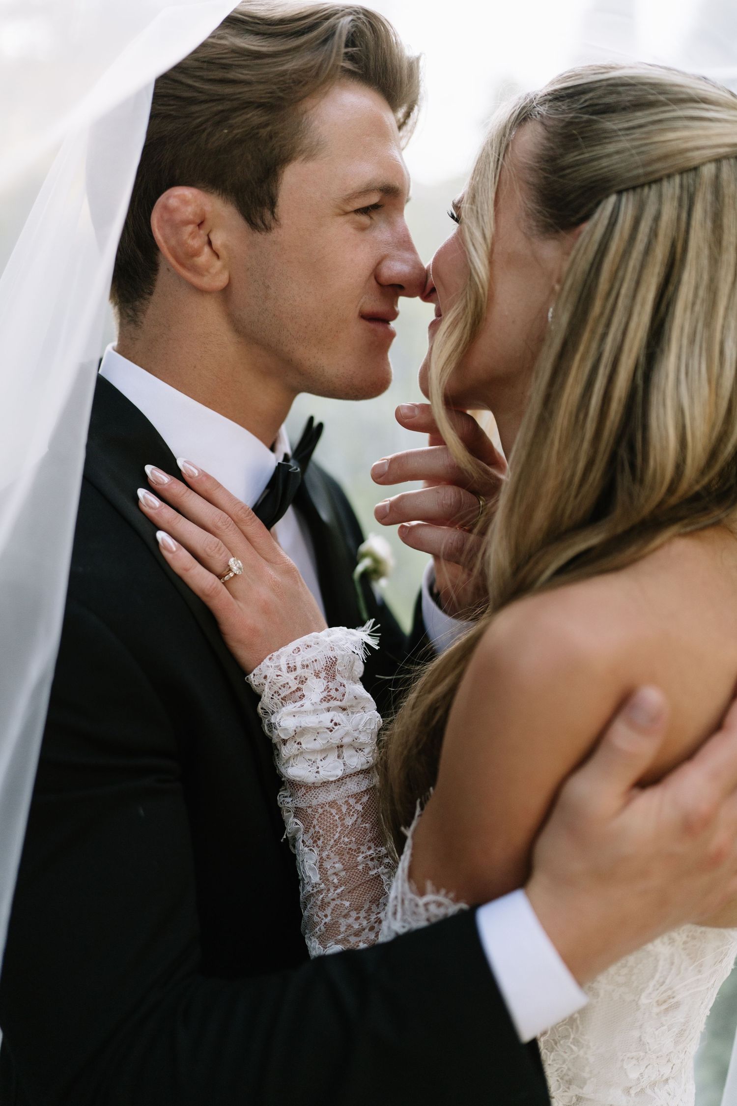 A bride and groom share an intimate moment, nearly kissing, as her lace sleeve and wedding ring are visible.