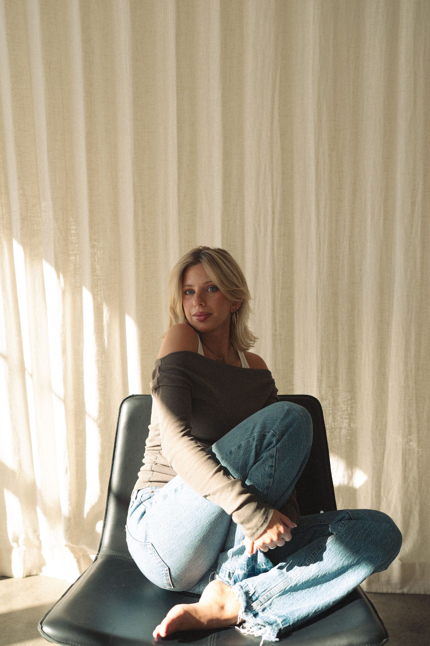 Person in black top and jeans sitting casually on dark chair against light curtains with dramatic lighting.