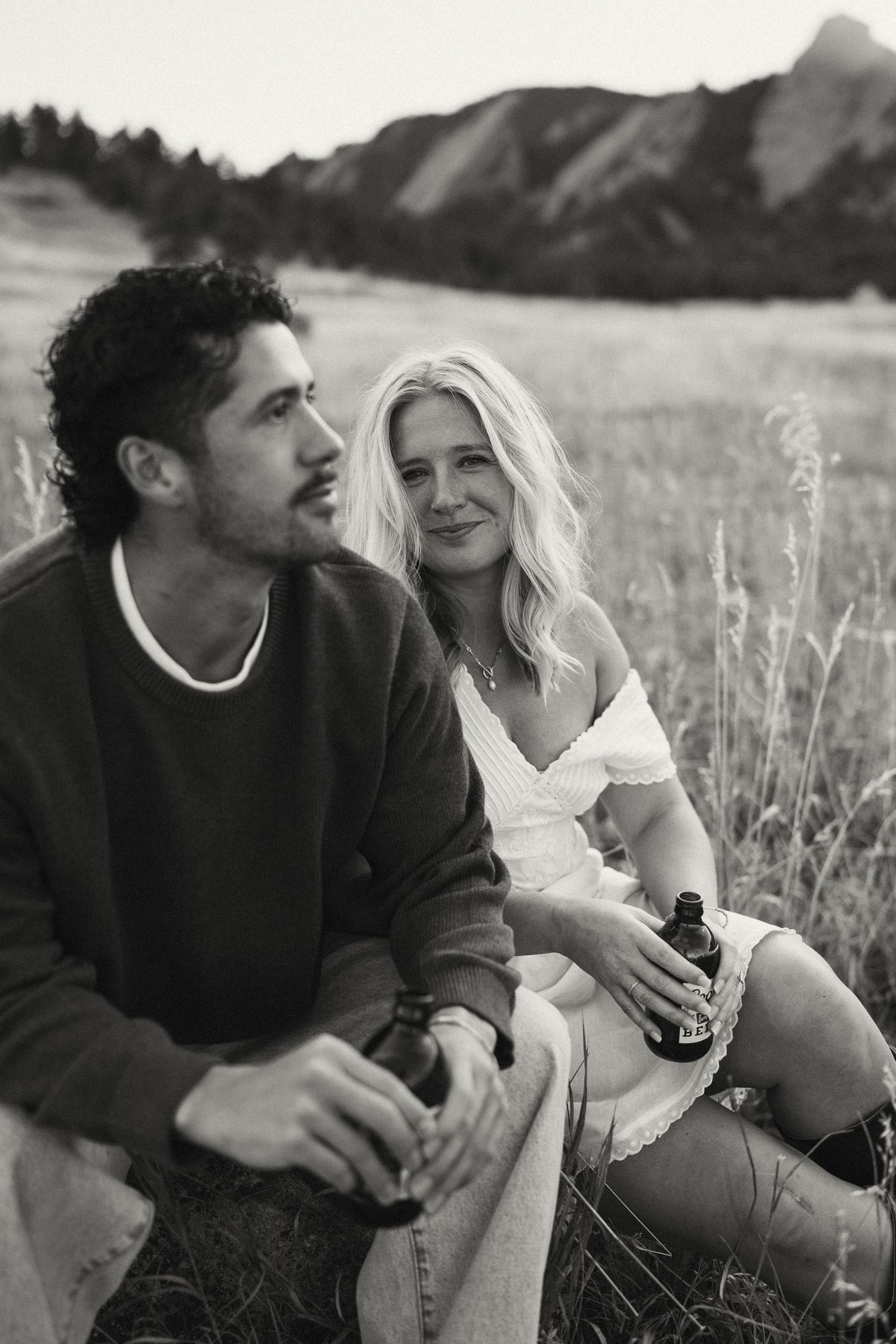 Couple sitting together in a grassy field sharing drinks and laughs at sunset.