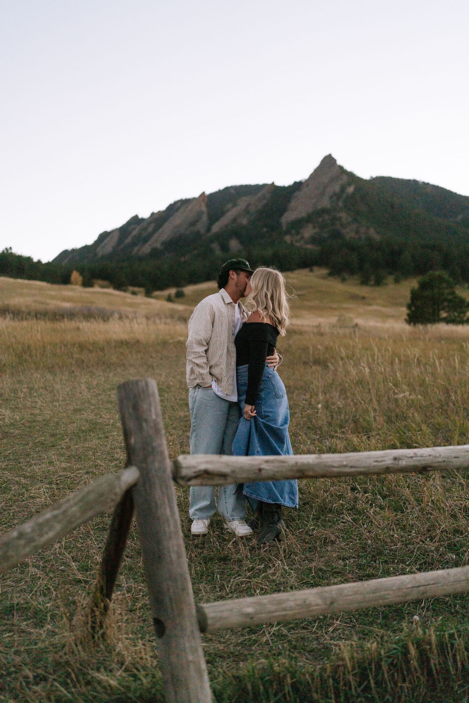 Couple enjoys tender moments near wooden fence with mountain backdrop.