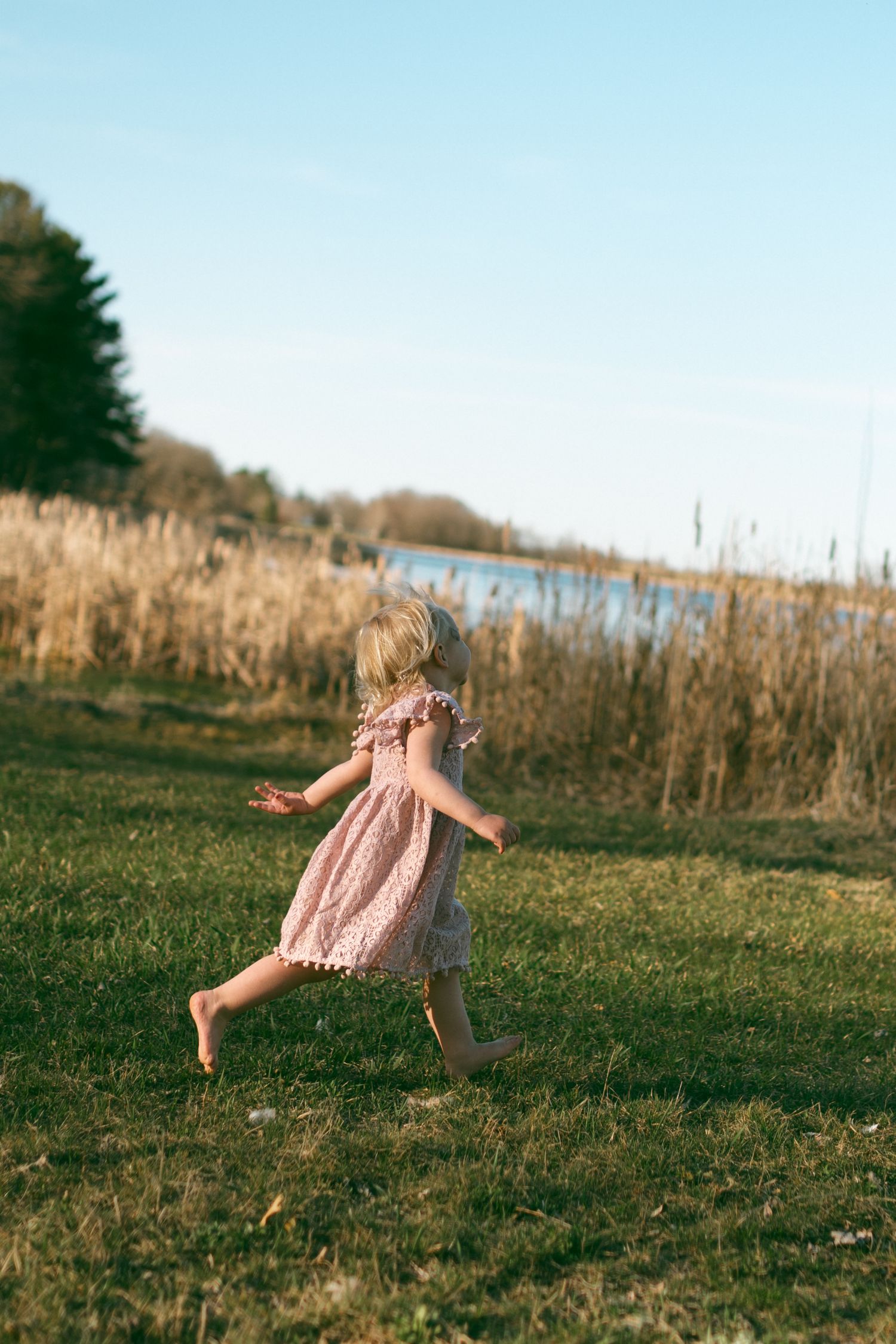 Young child in pink dress running freely through a grassy field near a lake.