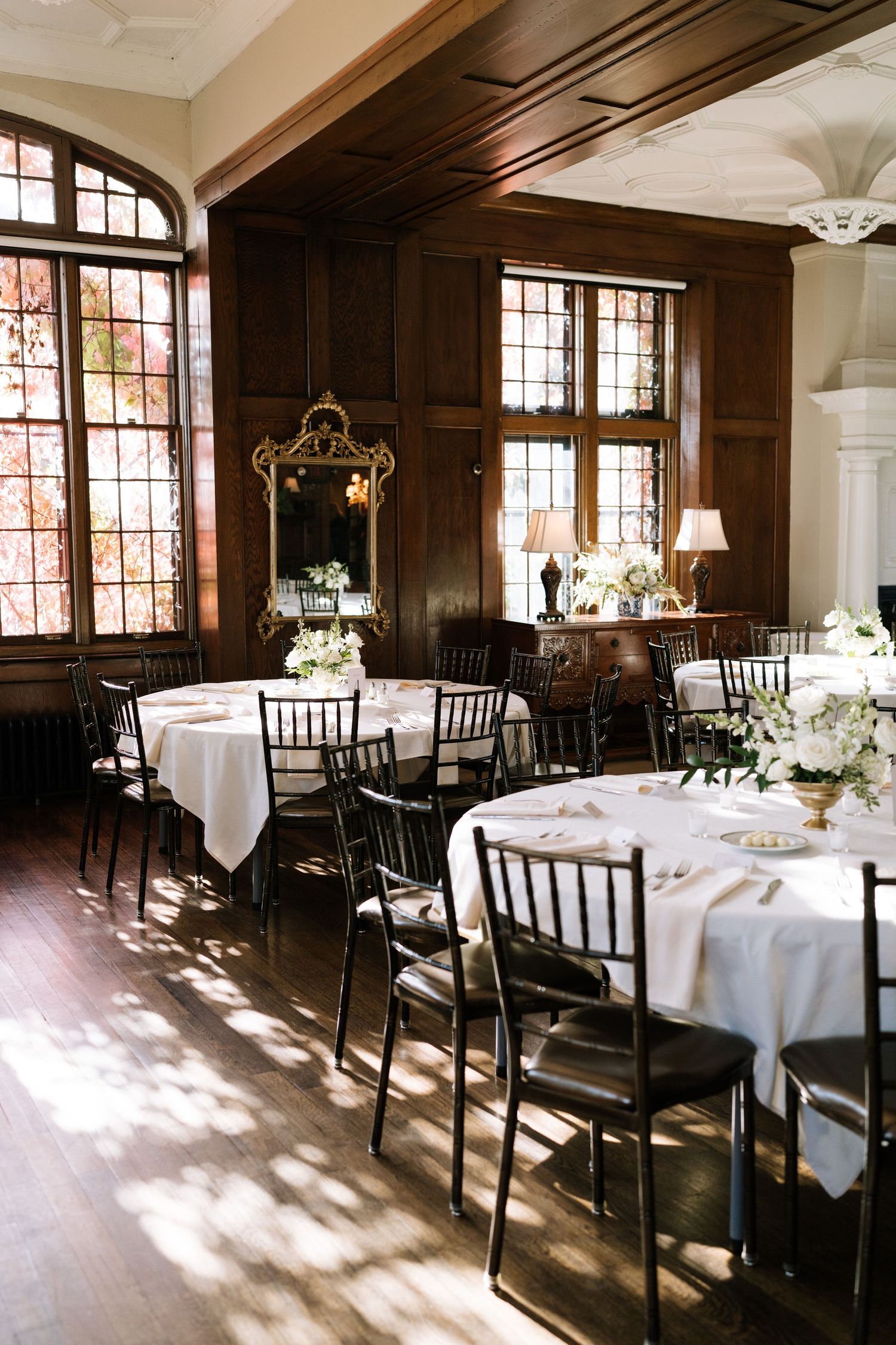 Elegant dining room with dark wood paneling, white tablecloths, and natural light streaming through windows.
